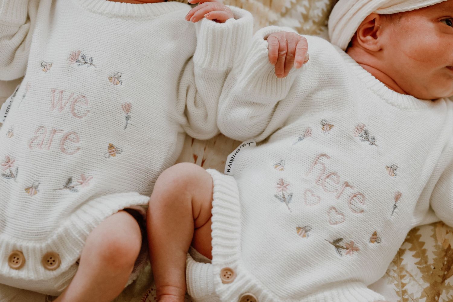Twin newborns in matching cream knit outfits reading we are here lying side by side.