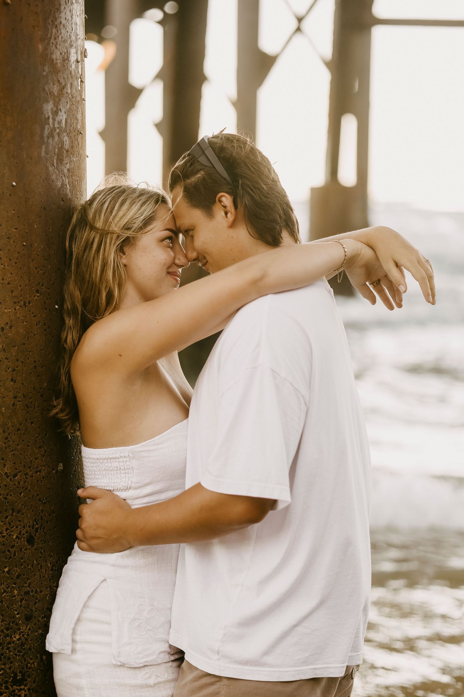 Young couple sharing a romantic moment leaning against a pier post at sunset beach.