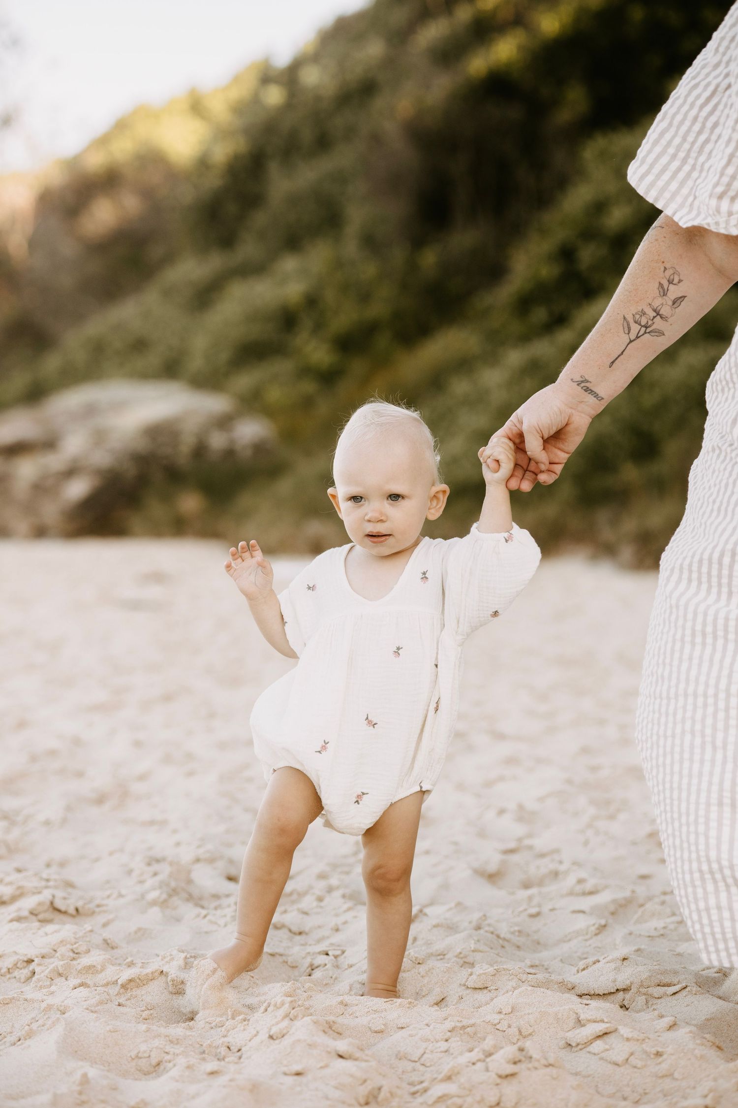 Baby in white romper taking first steps on sandy beach while holding parent's tattooed hand.