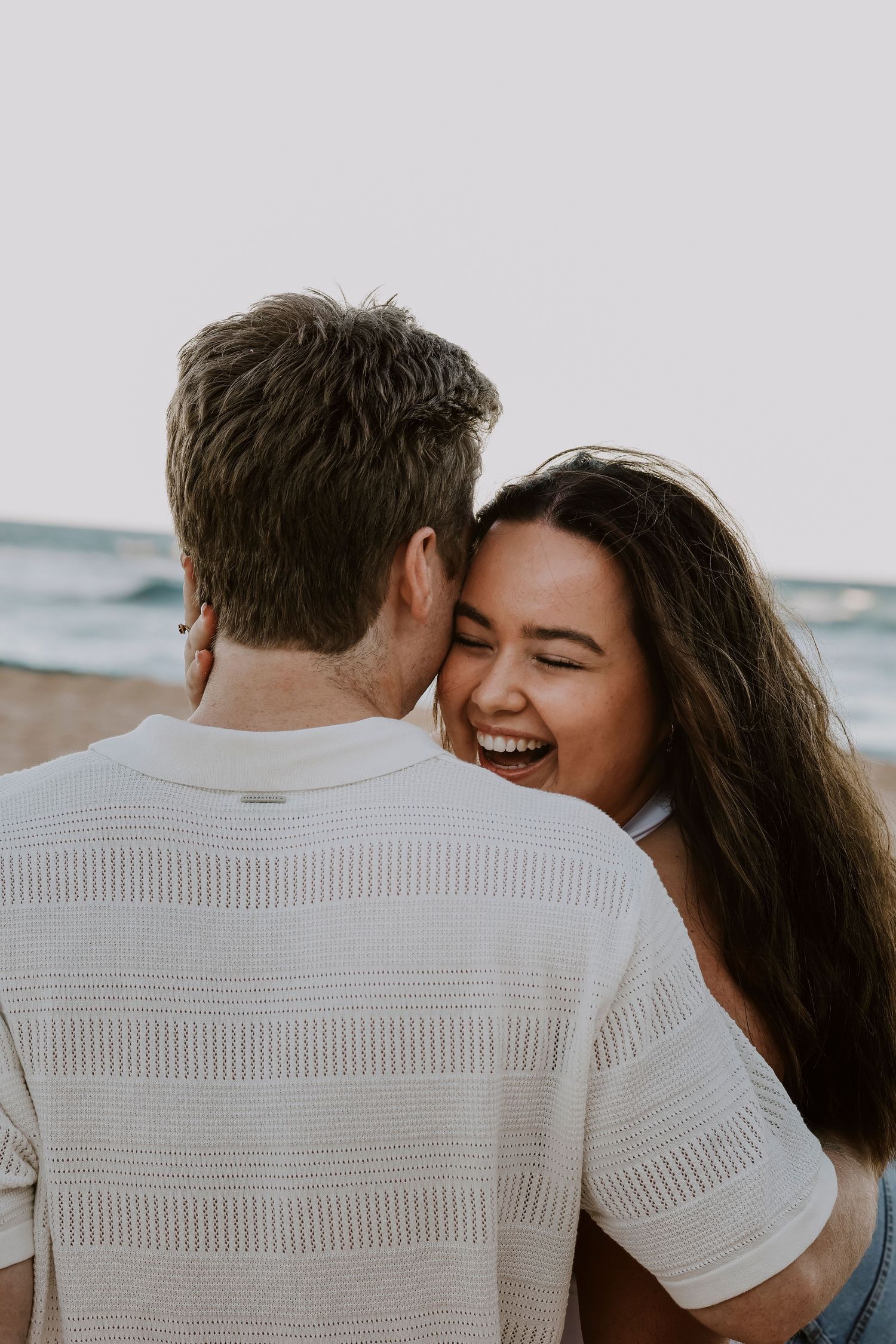 Couple sharing a joyful moment on the beach, woman laughing as man whispers in her ear.