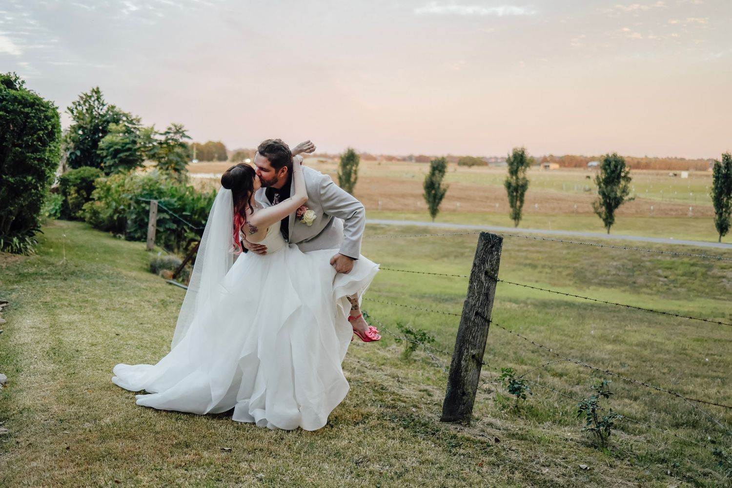 Groom playfully dips bride in a lush green countryside setting at sunset.