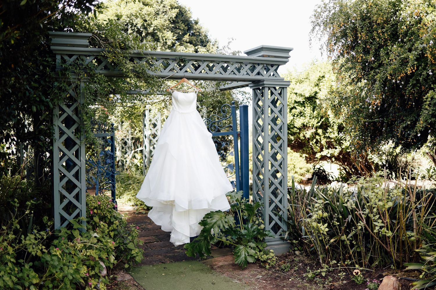 White wedding dress hanging on a blue garden trellis archway surrounded by lush greenery.