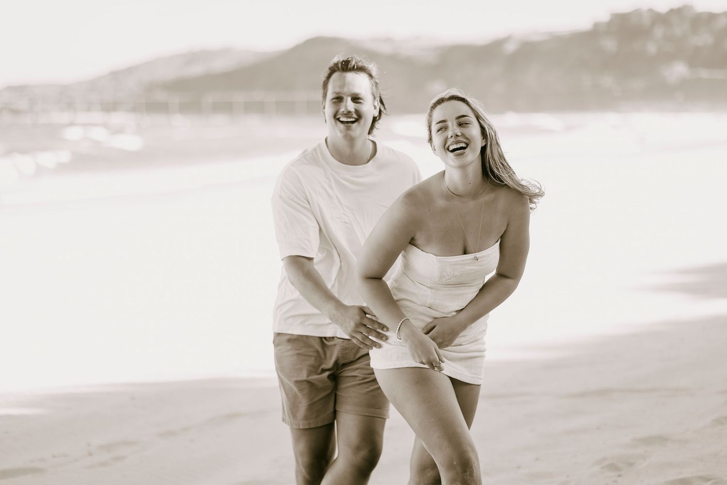Happy couple laughing and walking together on a sandy beach in black and white photo.