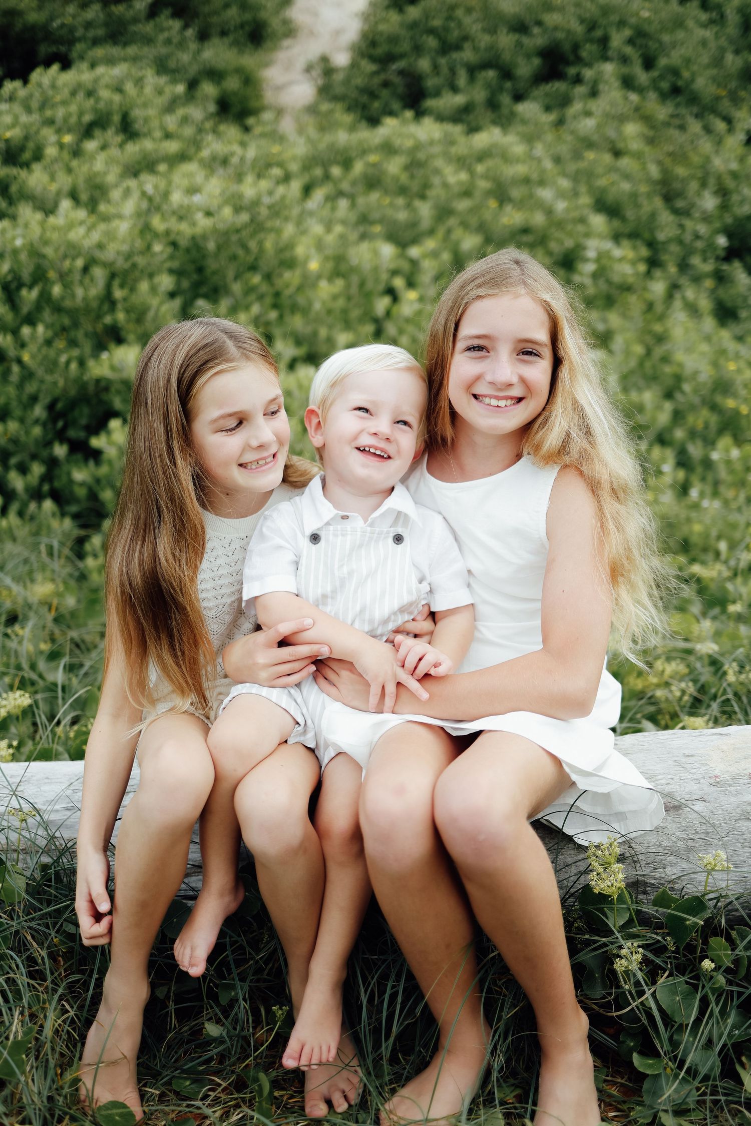 Three happy children sitting on a log outdoors, two girls with long hair and a toddler boy.