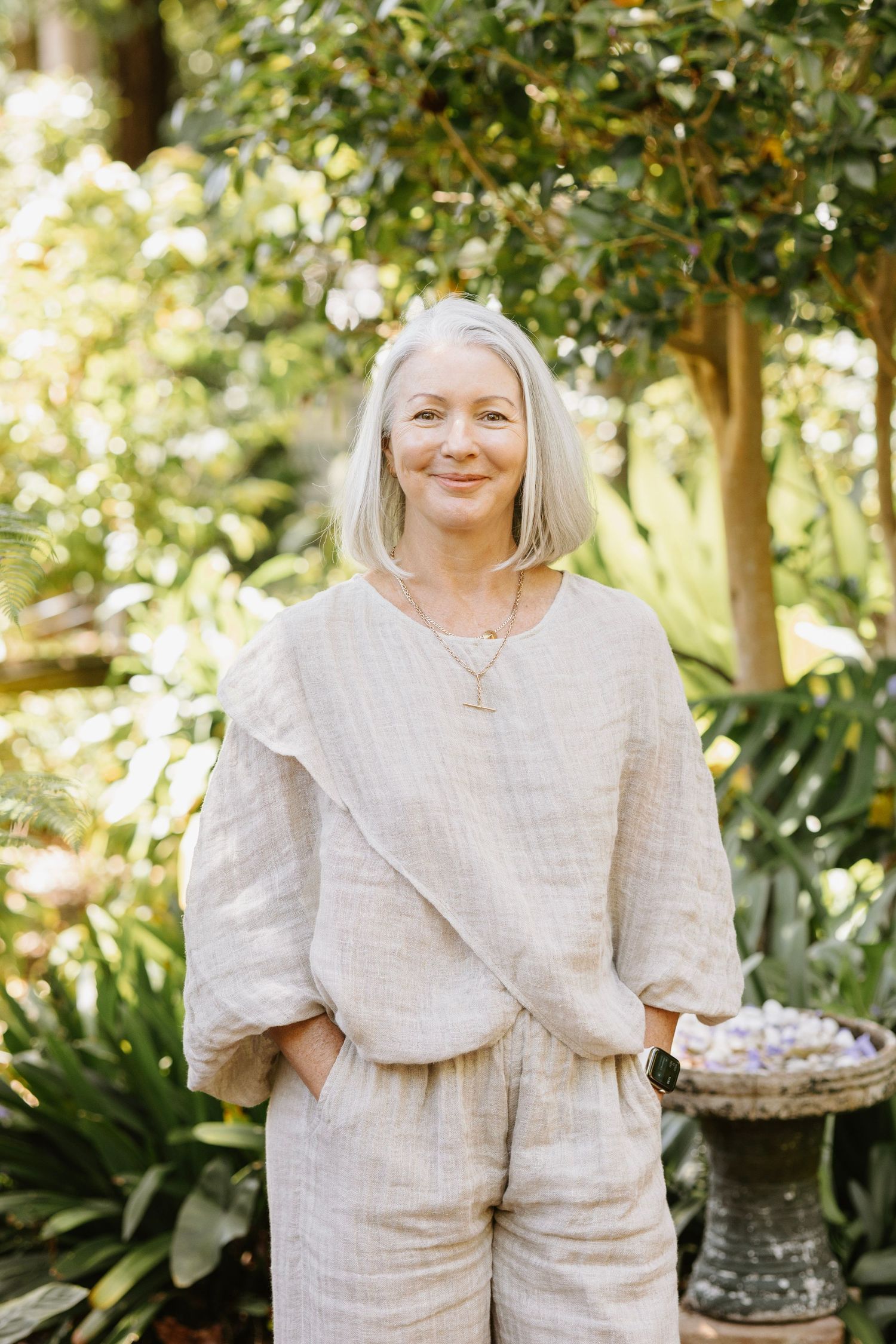 Smiling woman with silver hair in a beige linen outfit standing in a lush garden.