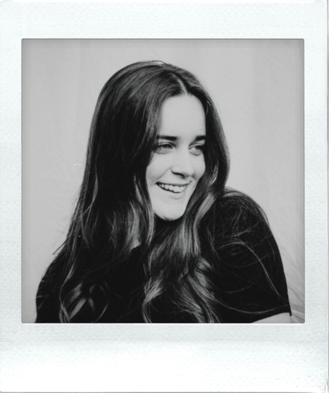 Black and white Polaroid photo of a cheerful young woman with long dark wavy hair smiling.