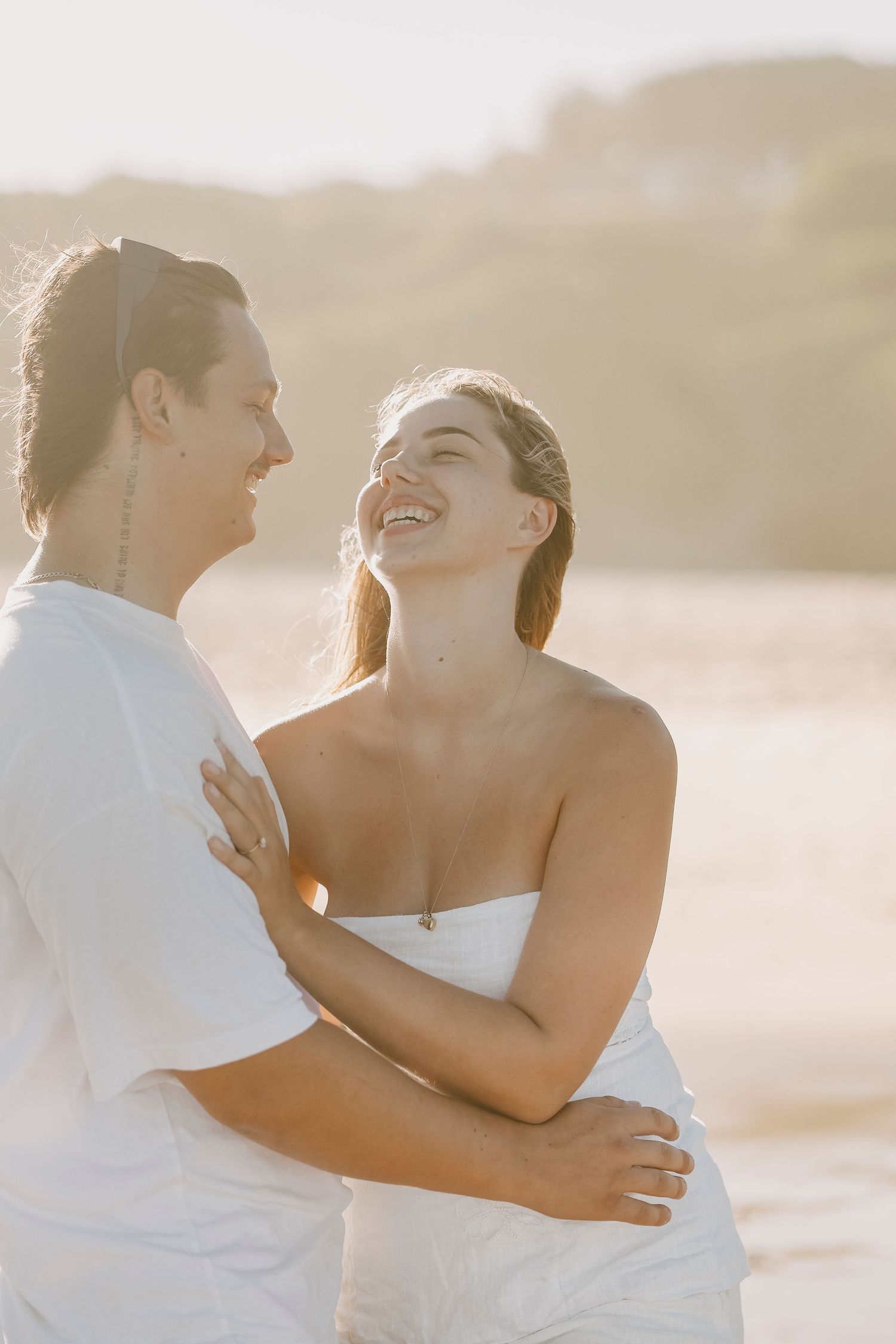 Glowing couple embracing and laughing on sunlit beach, woman in white top smiling up at partner.