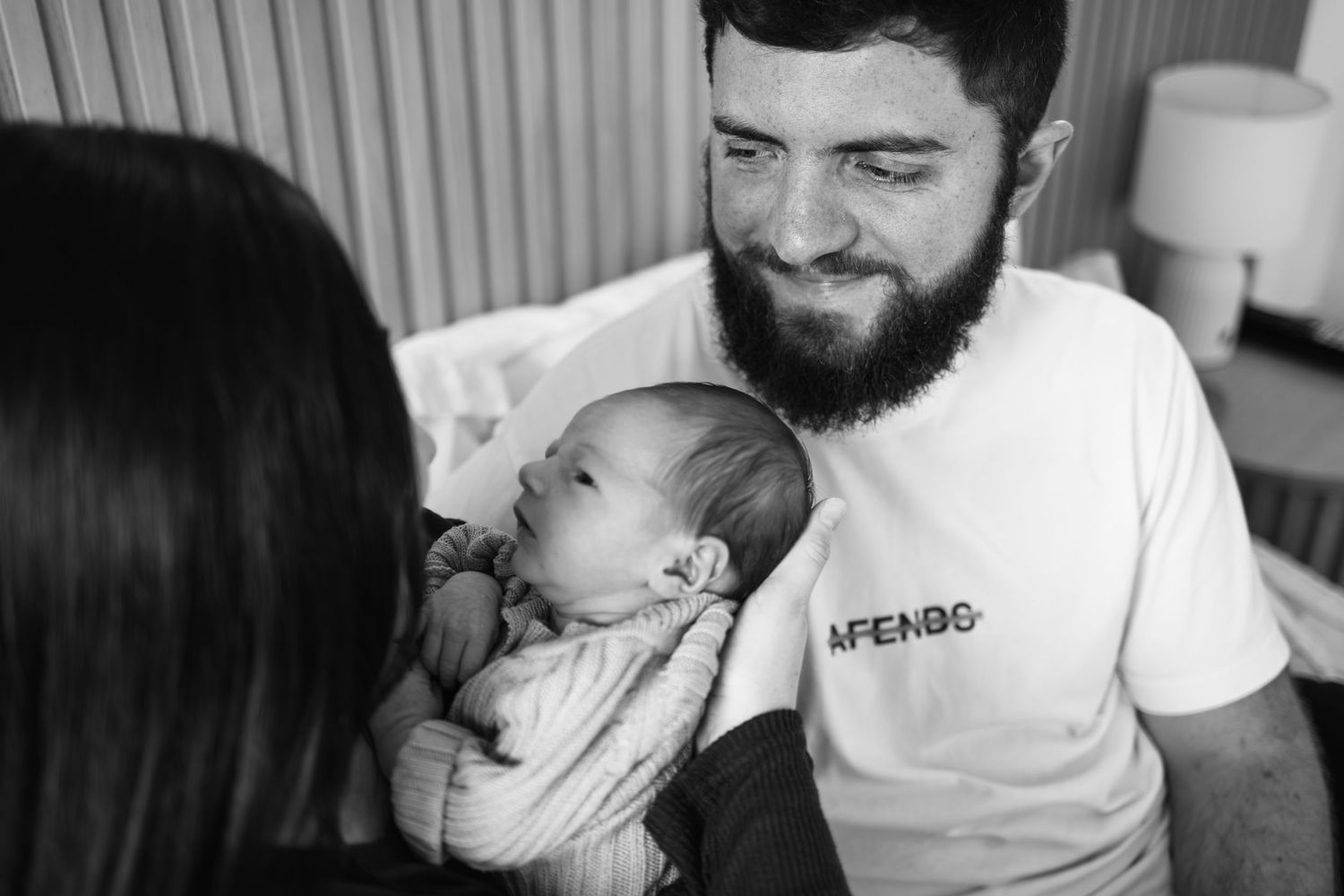Black and white photo of a bearded man in an Afends shirt tenderly holding a sleeping newborn baby.