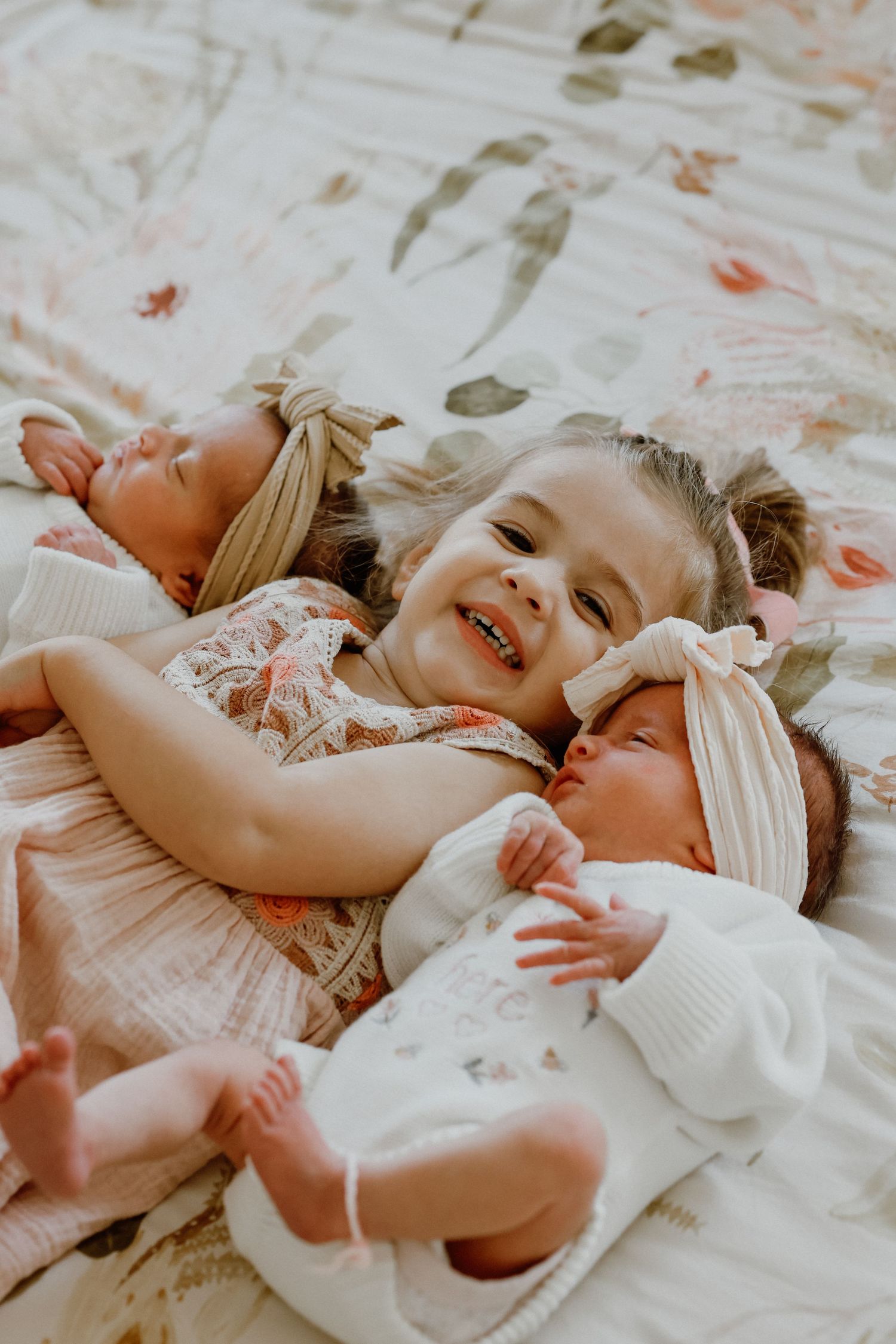 Excited young girl lying on a floral bed with two newborn twin babies on either side.