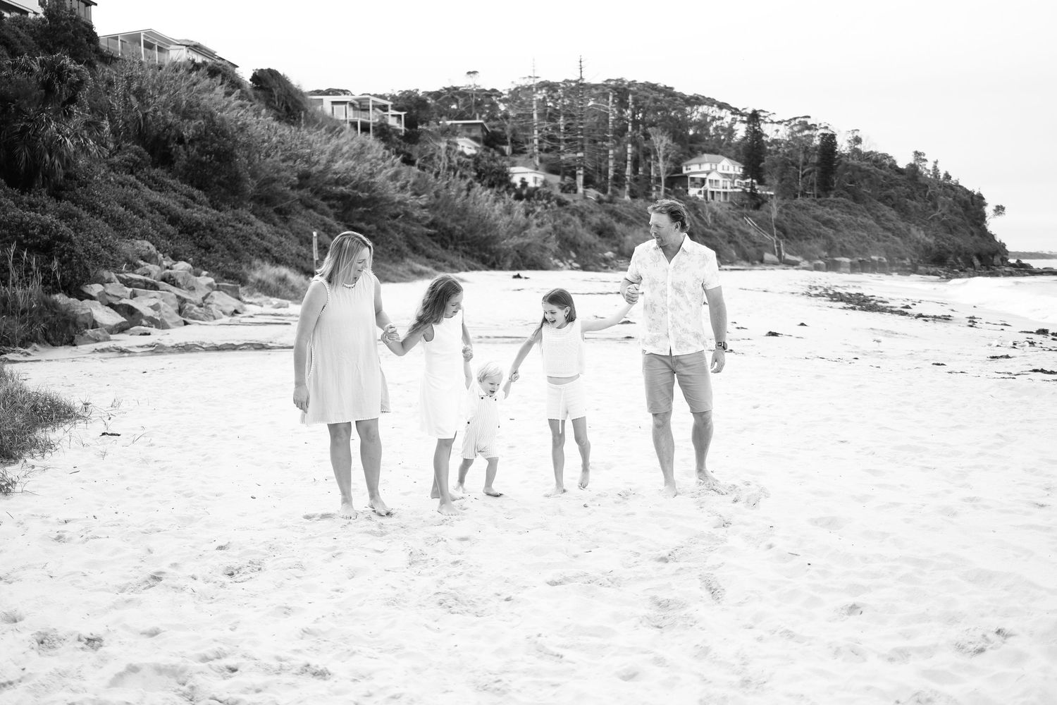 Black and white photo of a family of four walking hand in hand on a beach.