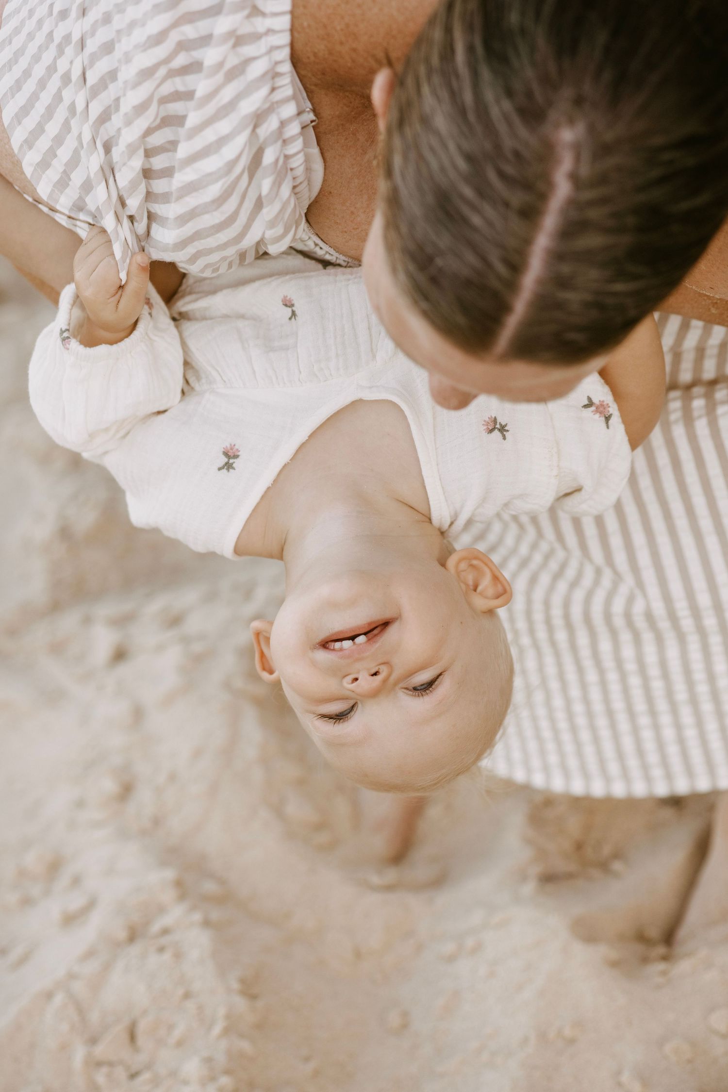 Mother holding smiling baby upside down on a sandy beach in warm natural light.