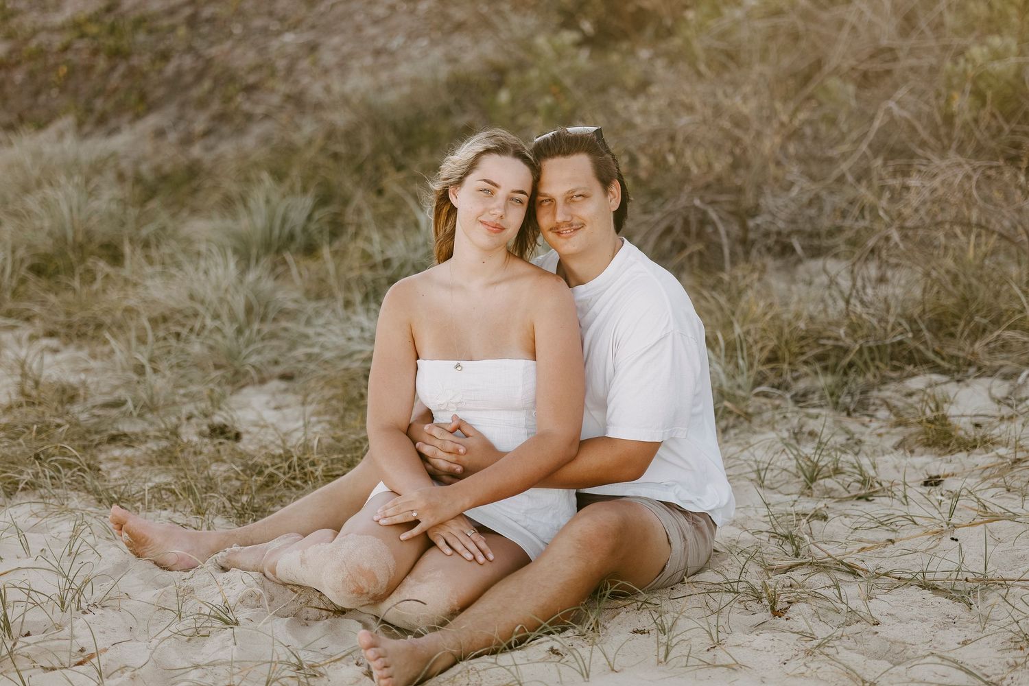 Happy couple sitting together on sandy dunes, woman leaning back into mans arms smiling warmly.