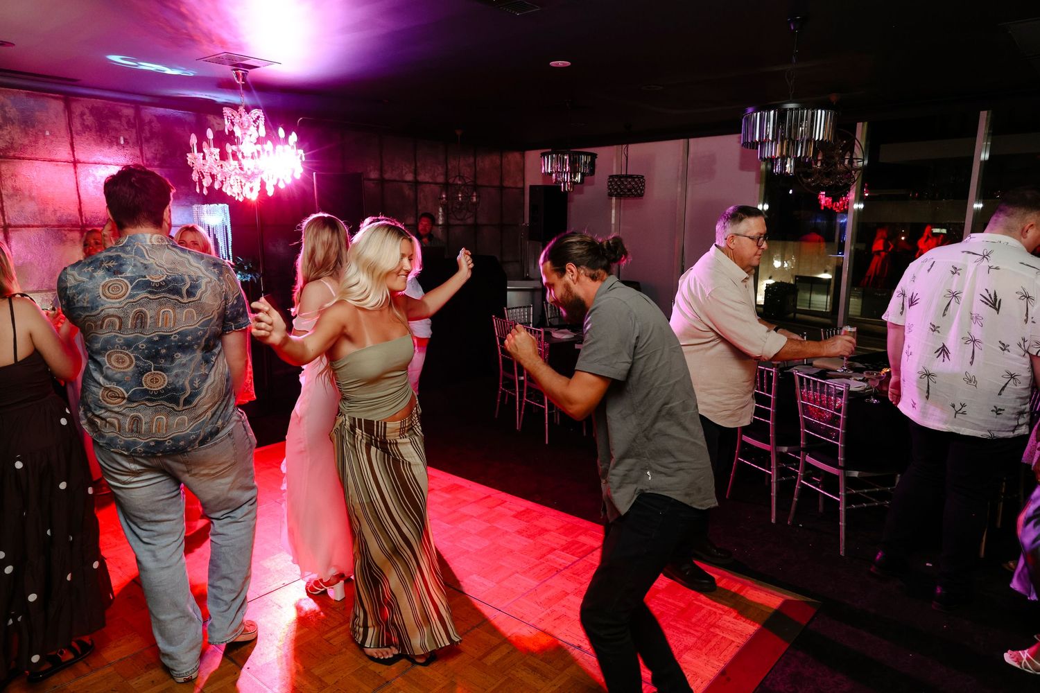 Guests dancing together at an elegant evening reception with chandeliers and warm pink lighting.