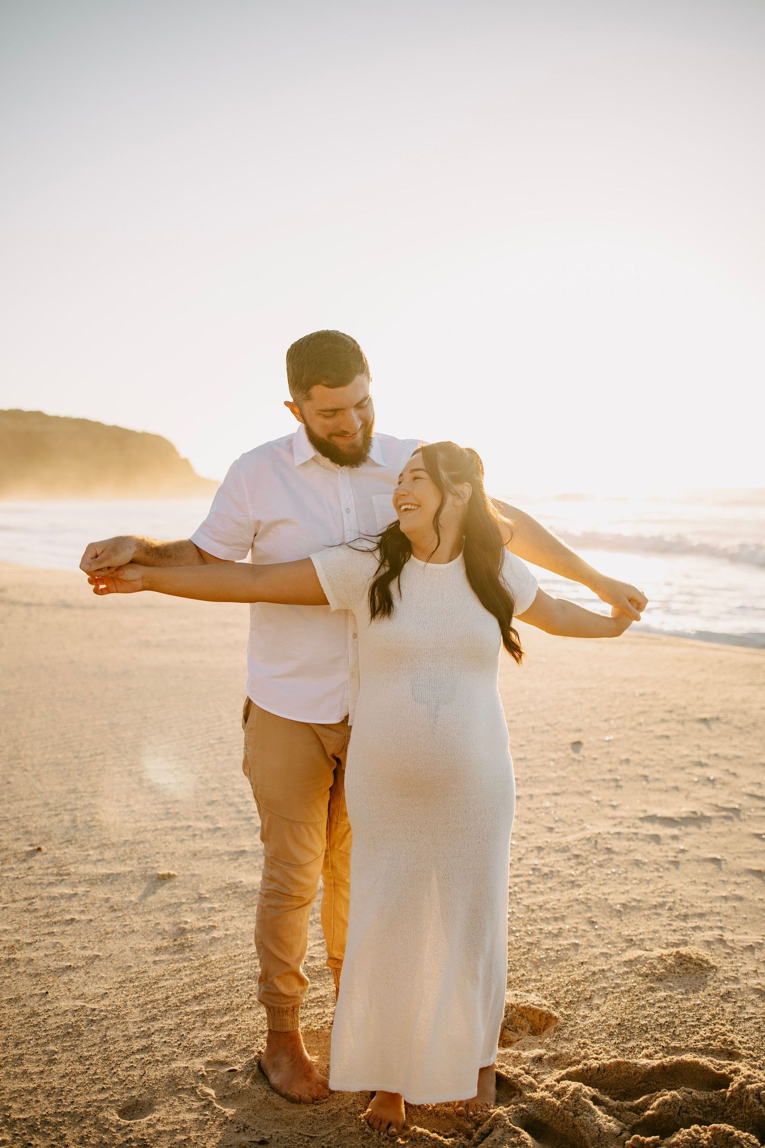 A happy couple dances together on a sandy beach at sunset, woman in a white maternity dress.
