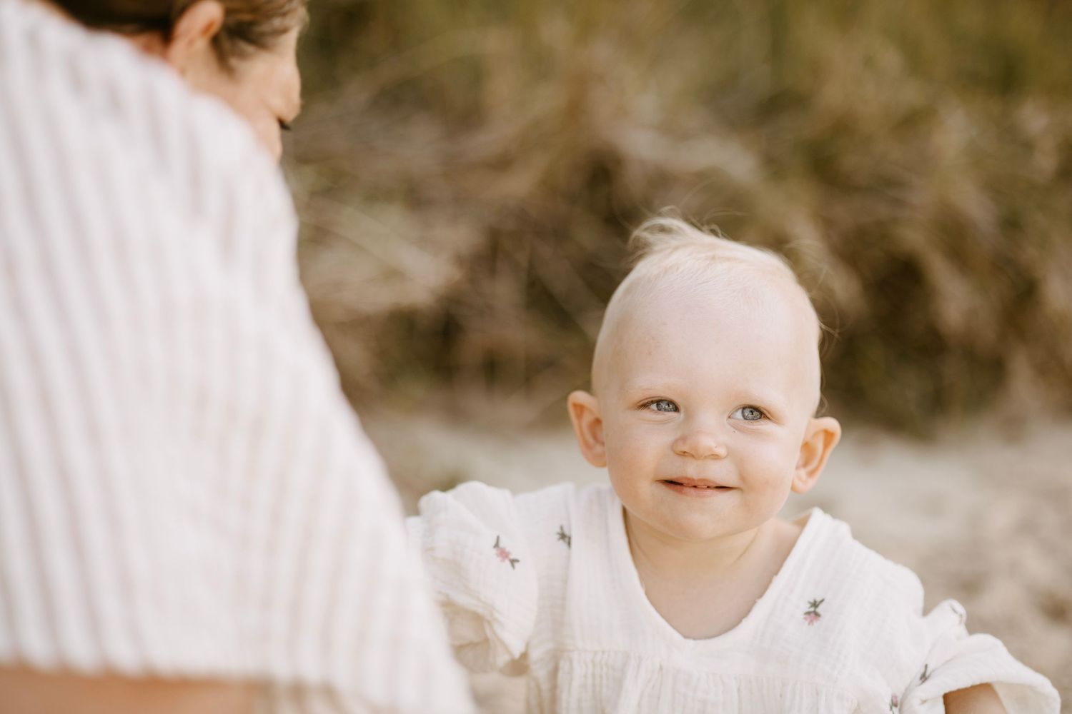 Smiling blonde baby in white floral romper looking up at parent on sunny beach.