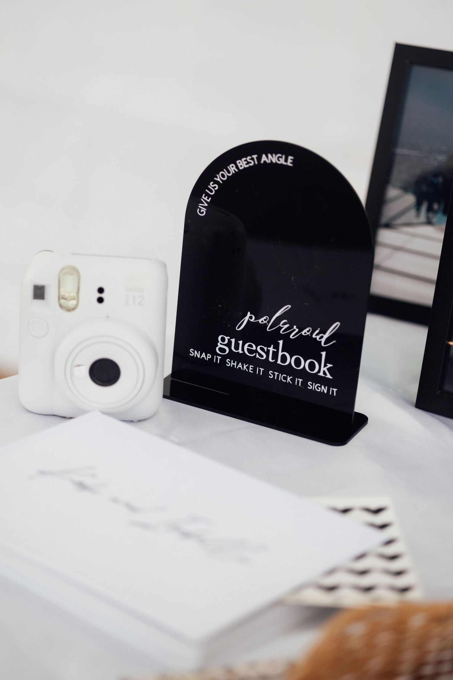 Polaroid guestbook station with Instax camera and black acrylic sign at a wedding or party event.