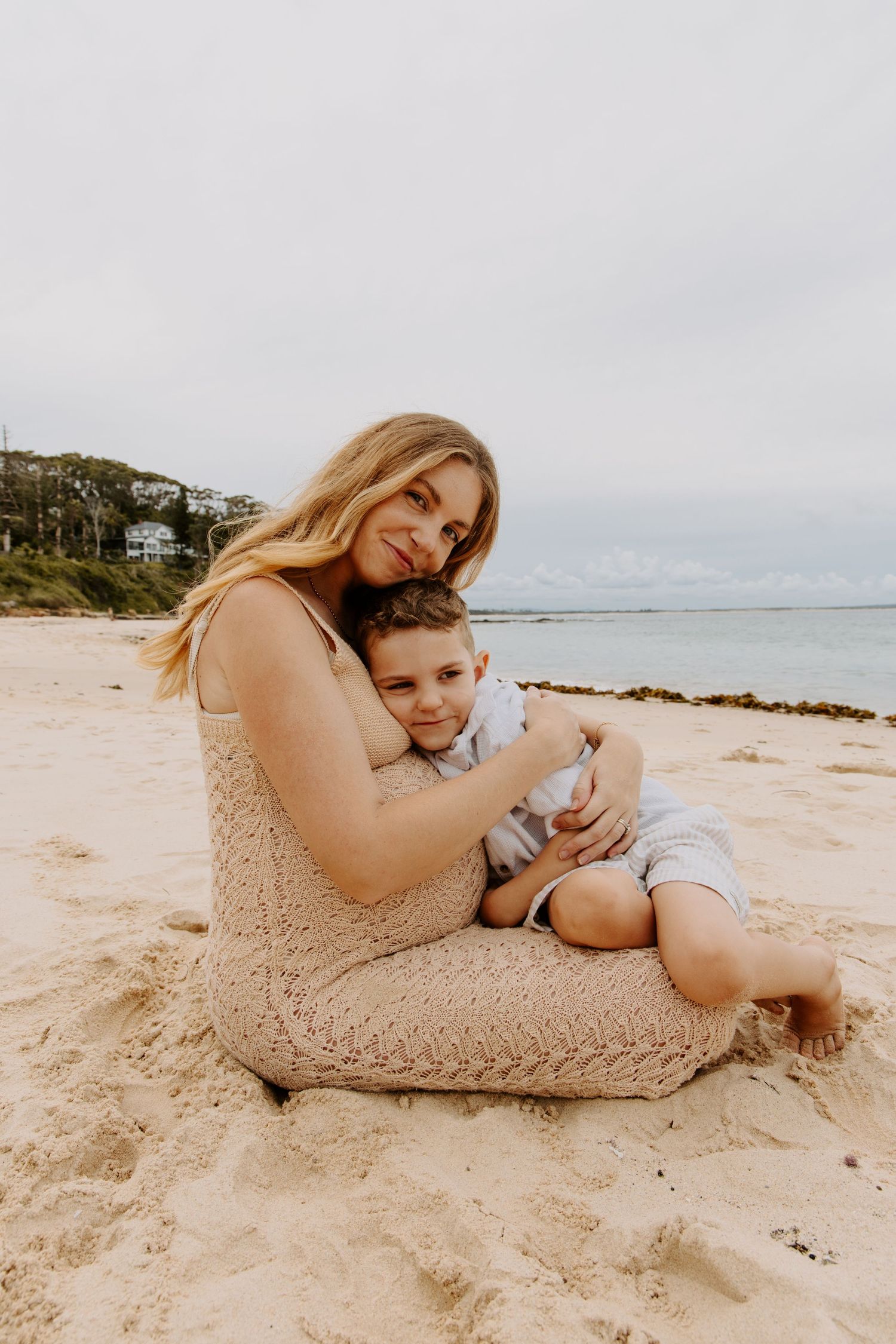 Mother in beige knit dress sitting on beach sand hugging young boy in white outfit.
