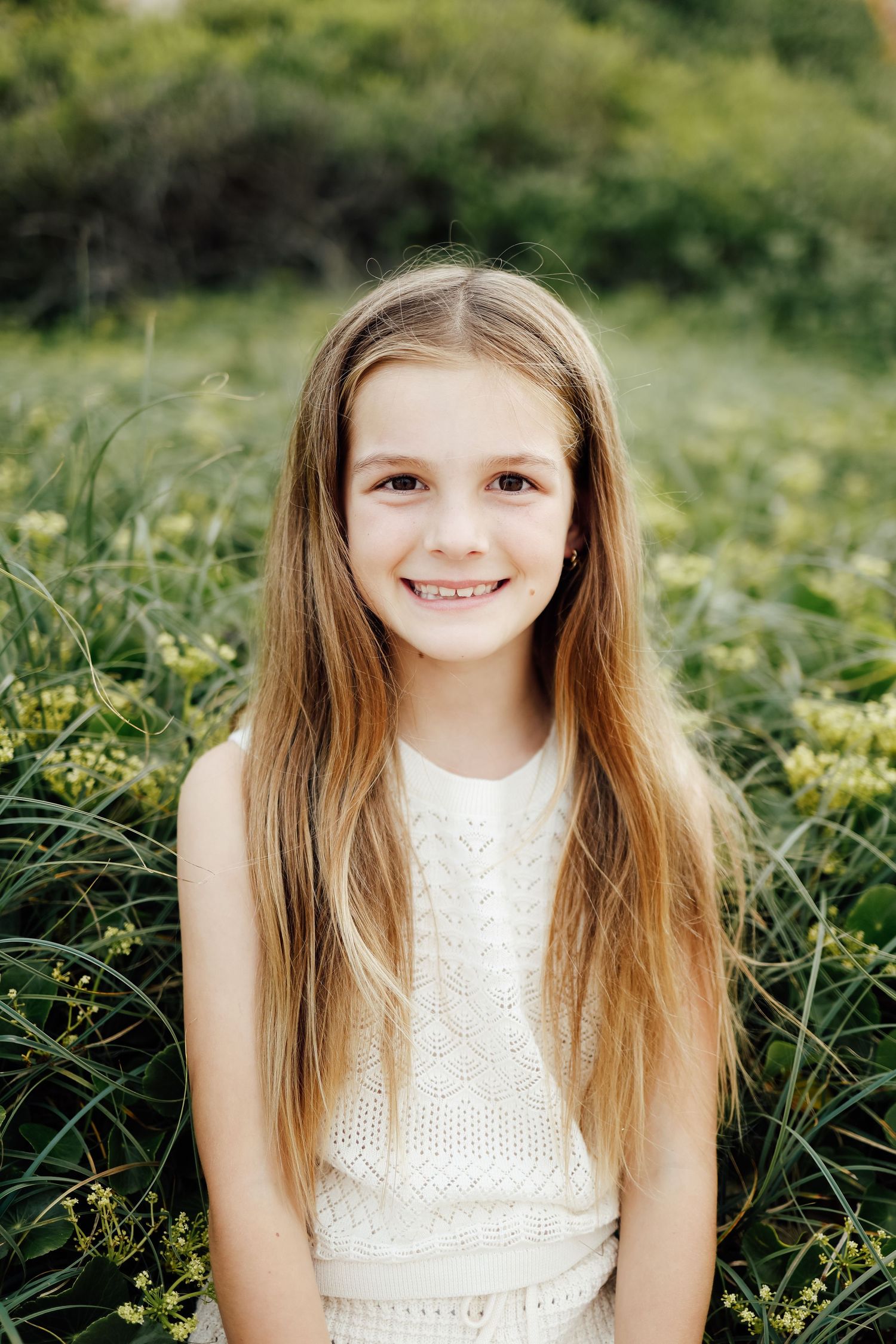 Smiling brunette girl with long hair in white top sitting among green wildflowers.
