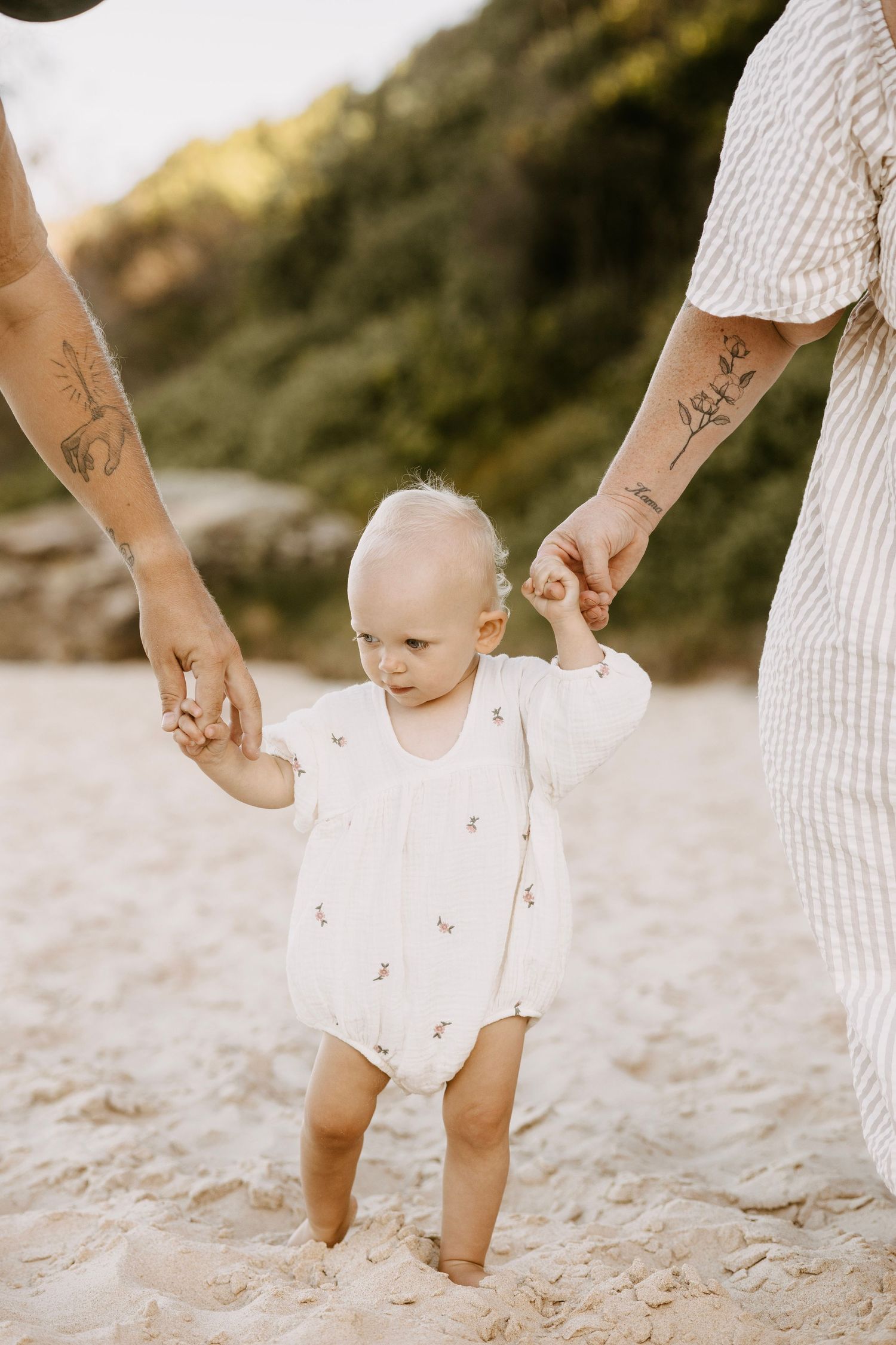 Baby in white romper learning to walk on sandy beach held by tattooed parents.