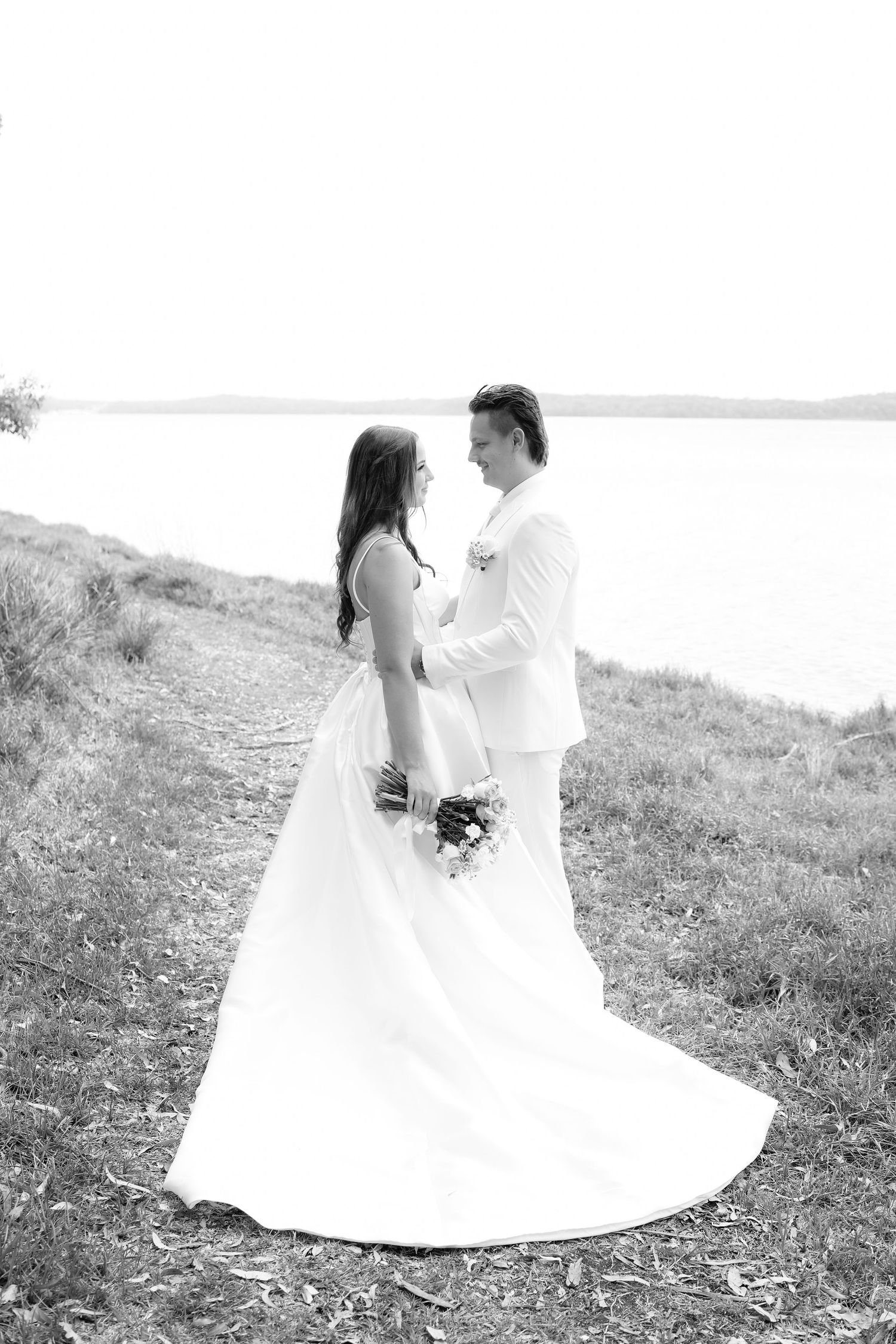 Black and white photo of a bride and groom holding hands by a lakeside, bride in flowing gown holding bouquet.