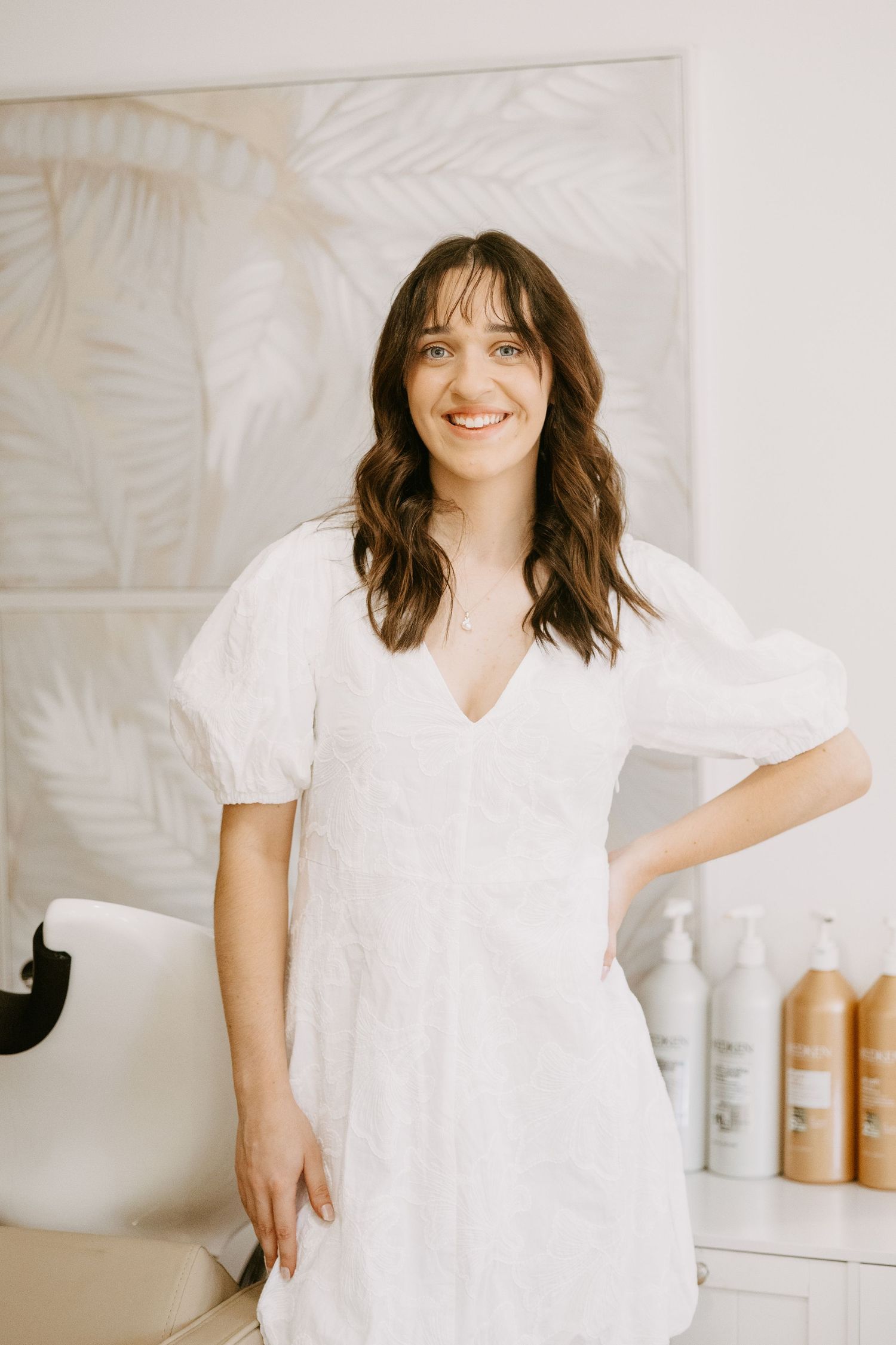 Smiling brunette woman in a white dress standing in a bright hair salon.