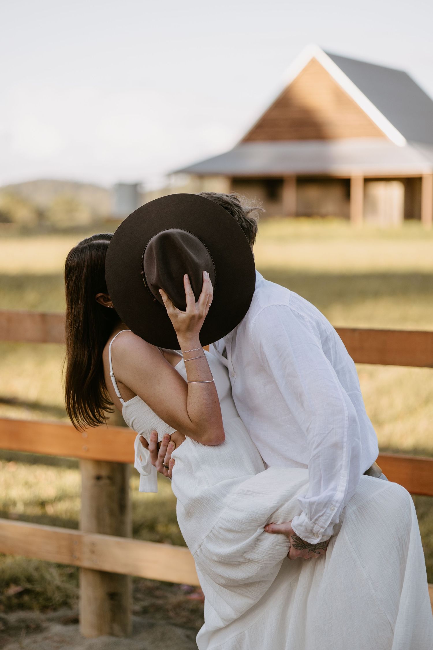 Couple sharing a kiss hidden behind a wide-brim cowboy hat leaning on a wooden farm fence.