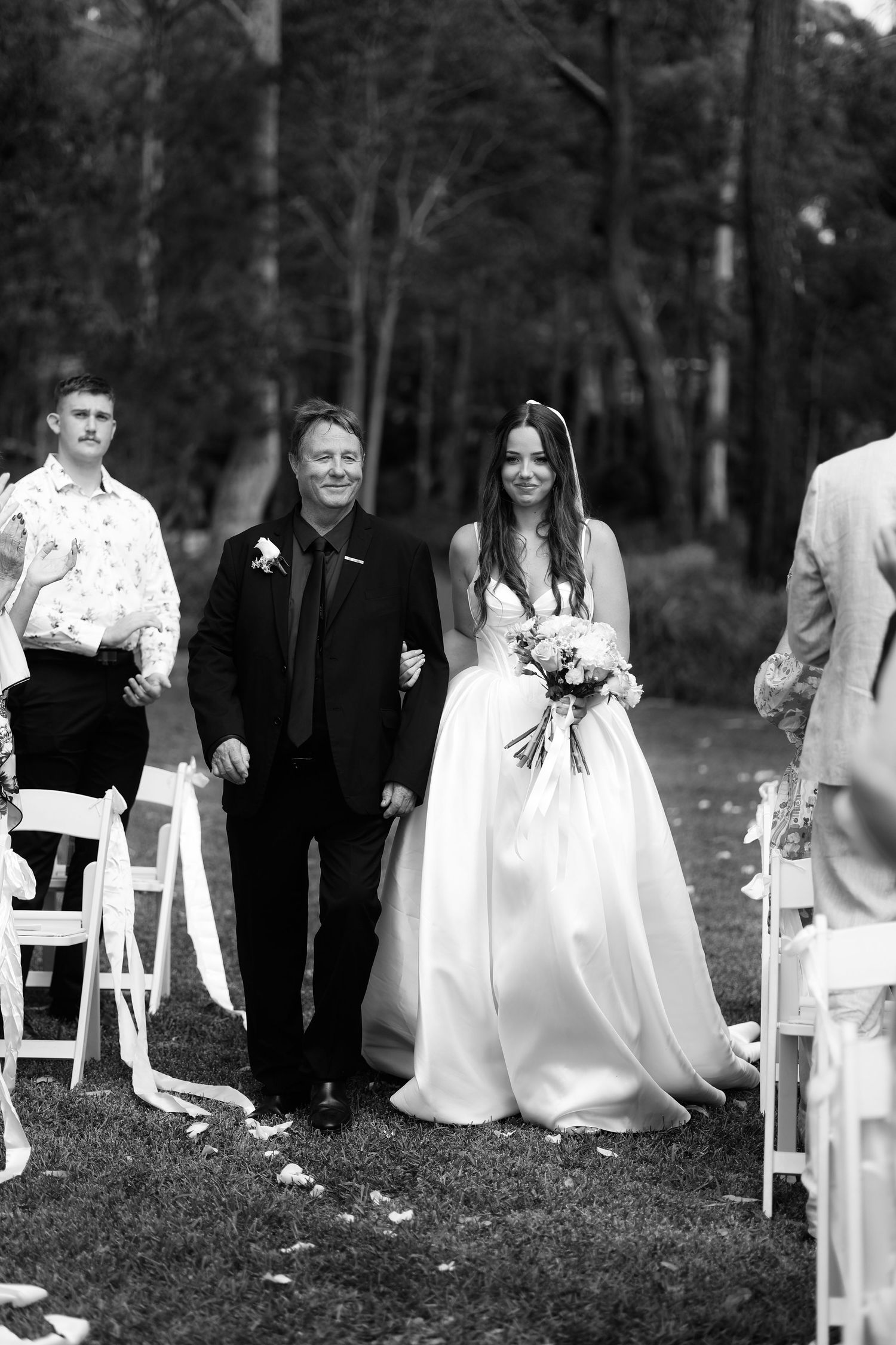 Bride walking down outdoor aisle with escort during wedding ceremony in black and white.