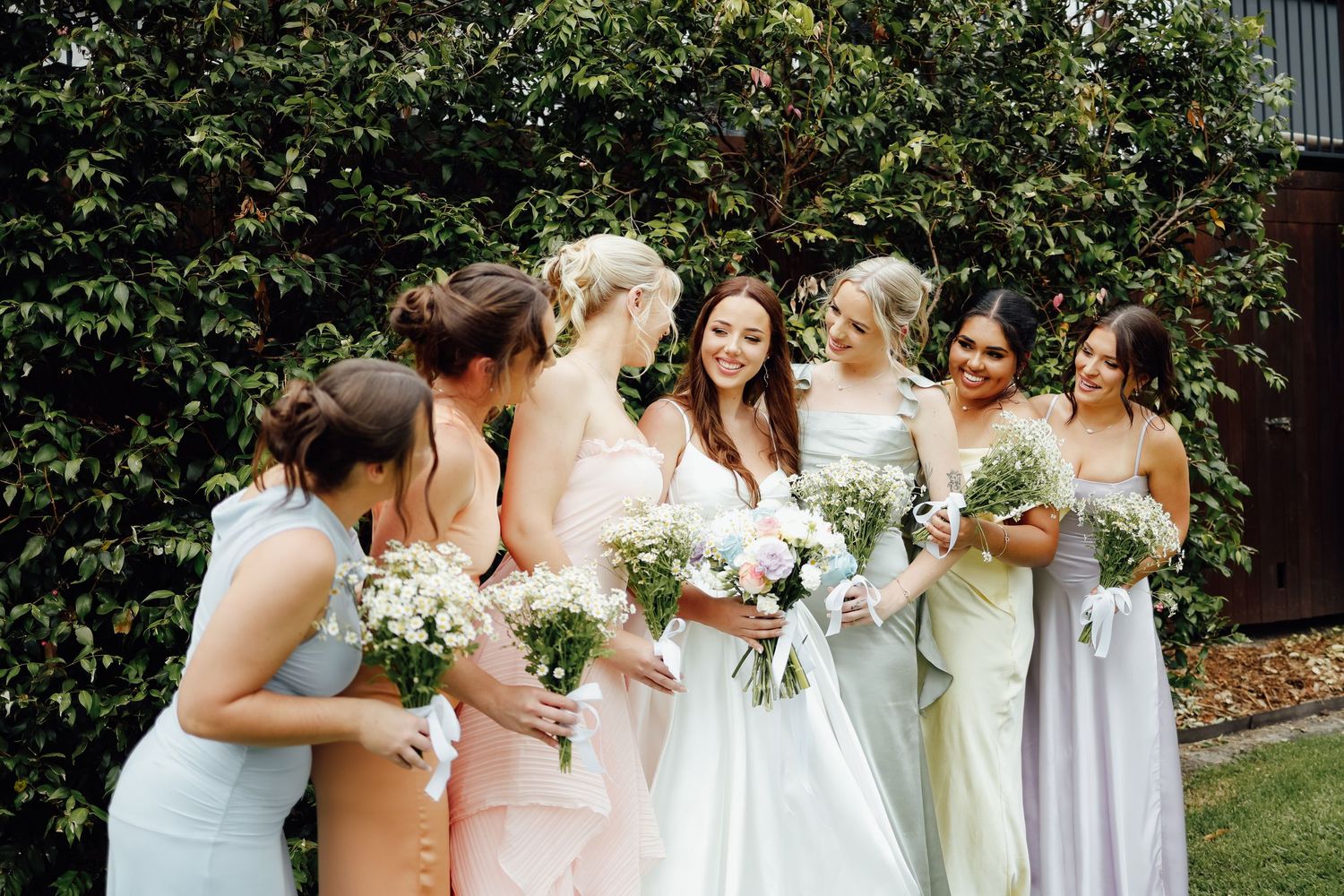 Bridesmaids in pastel dresses holding white bouquets laughing together outdoors.