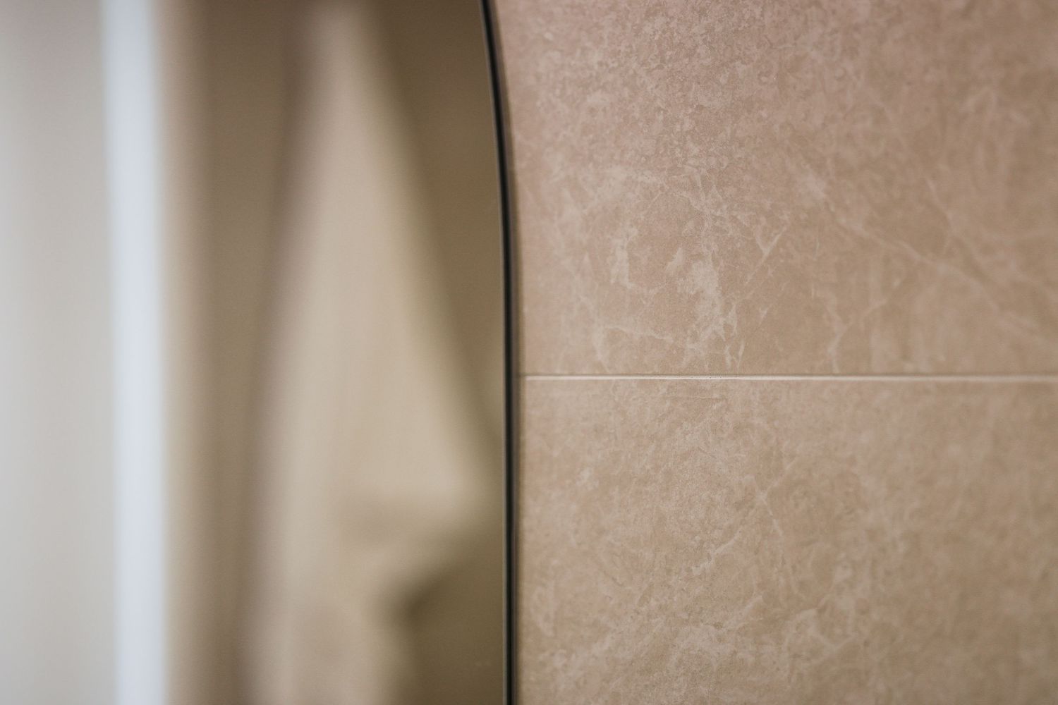 Close-up detail of a curved mirror edge against beige marble bathroom wall tiles.