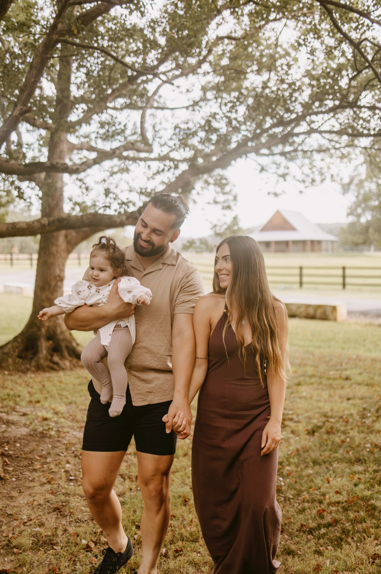Happy family with father holding baby girl and mother in brown dress walking under large tree.