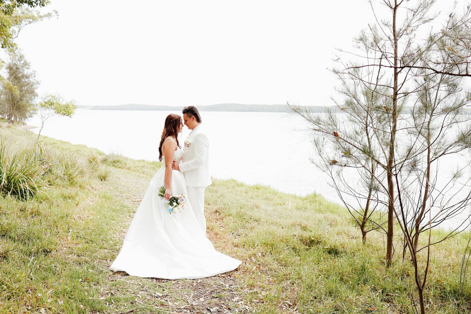 Bride and groom share romantic moment by serene waterfront surrounded by lush greenery on wedding day.
