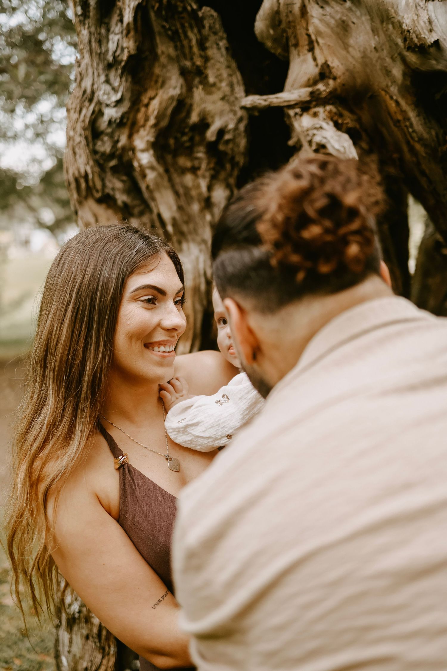 Smiling woman in brown dress gazes at partner holding baby near rustic tree outdoors.