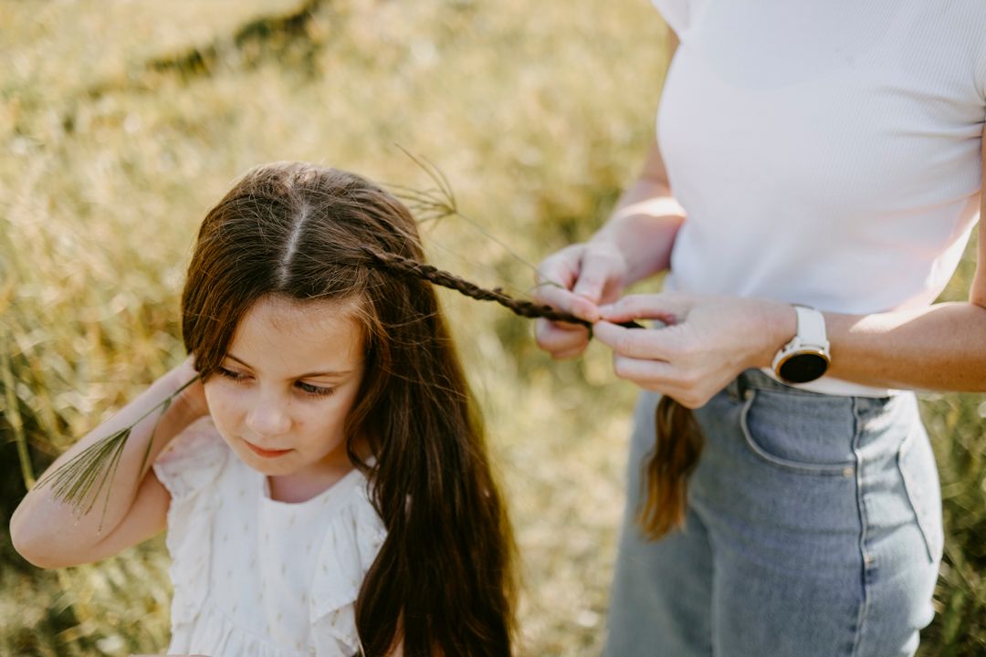 Adult braiding a young girl with long dark hair outdoors in a golden grass field.