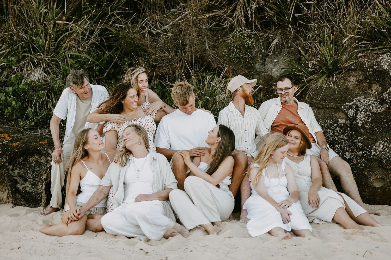Large group of friends in white outfits posing together on sandy beach.
