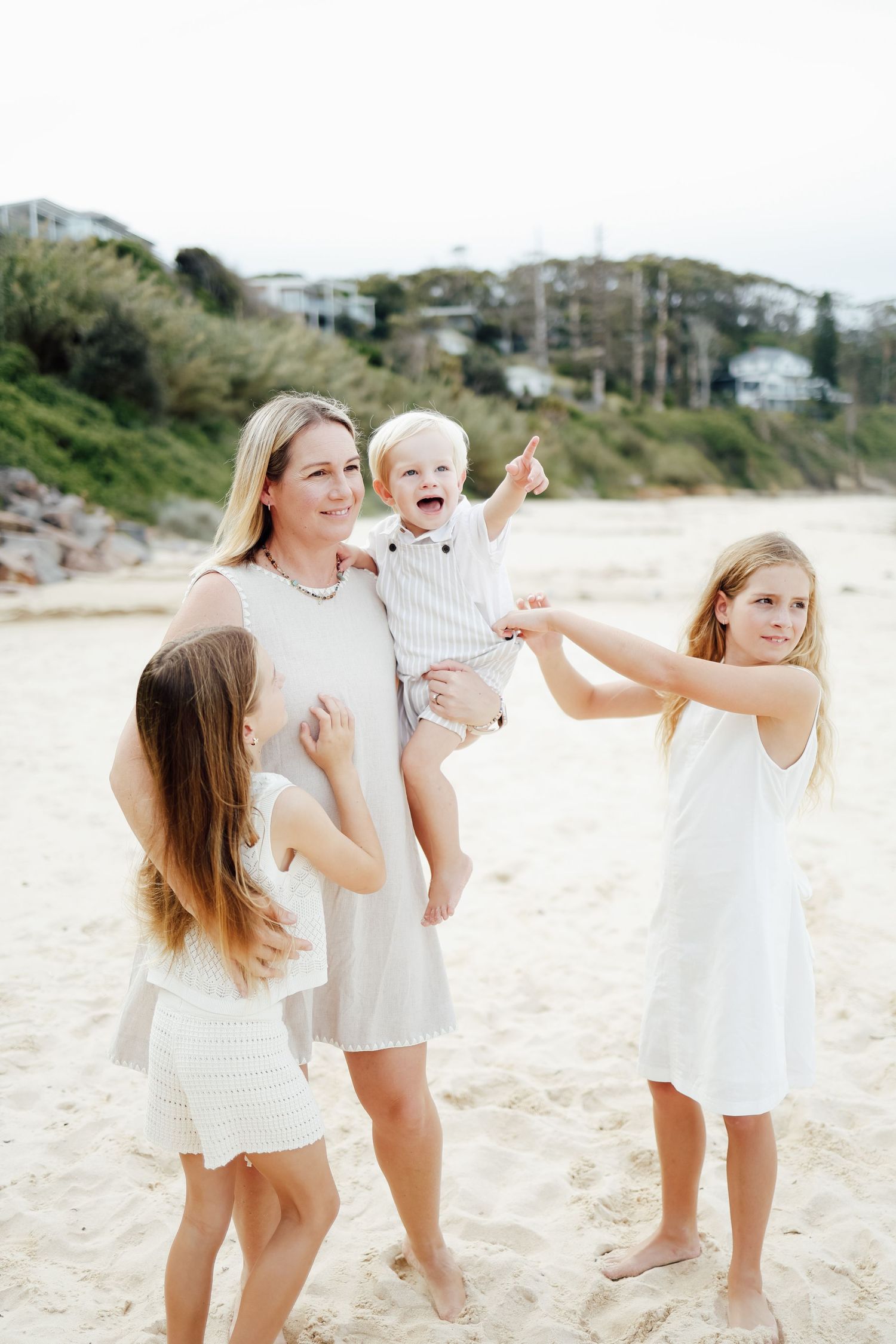 Mother in white dress holding laughing toddler boy on beach while two daughters play alongside.