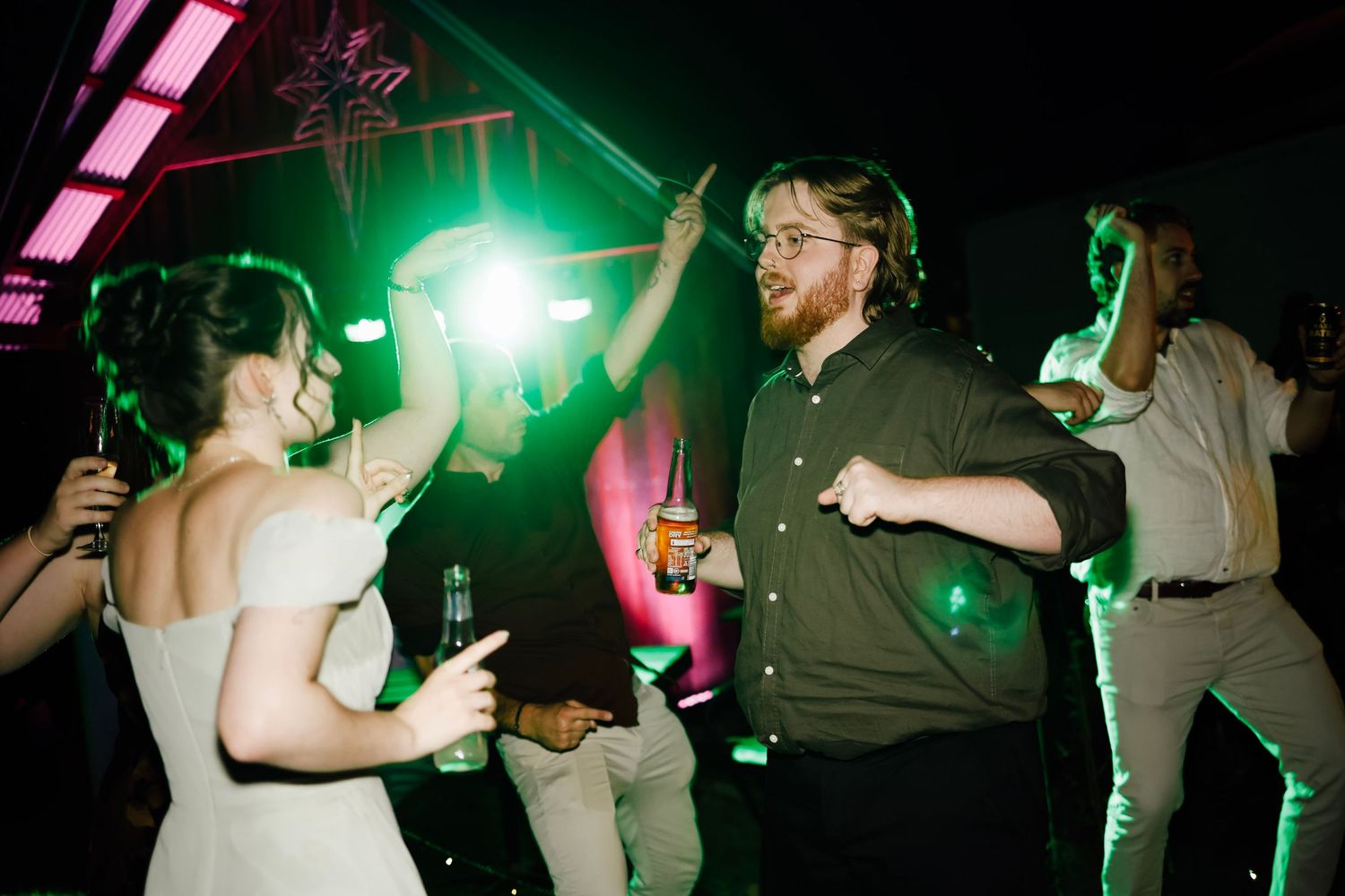 Bride and groom dancing joyfully at wedding reception under green stage lights with guests in background.