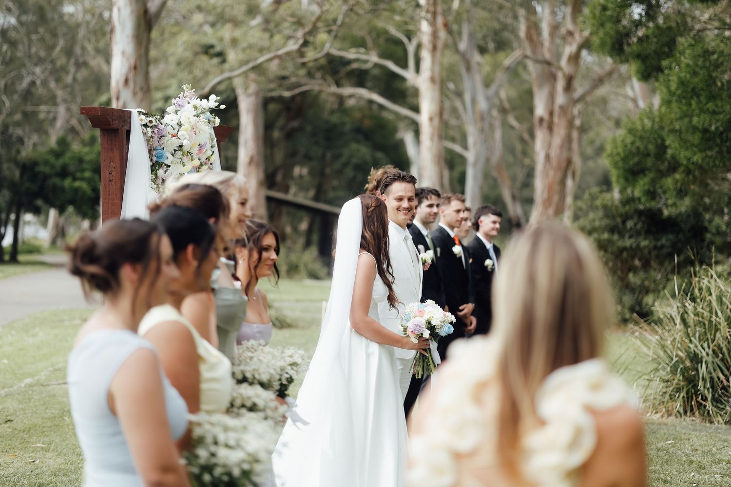 Bride in white gown smiles at bridesmaids during outdoor wedding ceremony surrounded by tall trees.