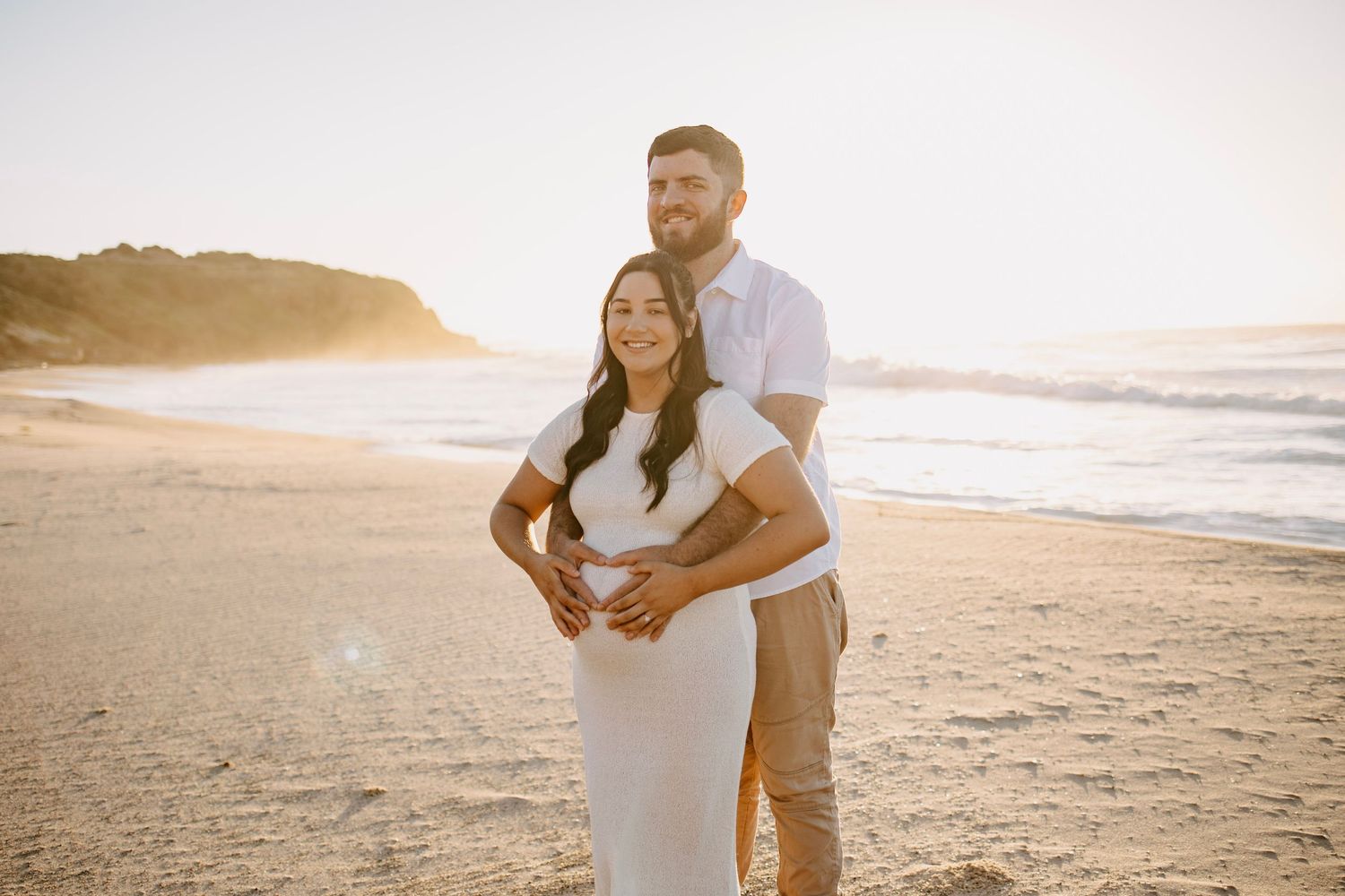 A couple sharing a tender moment on a sunny beach during a golden sunset maternity shoot.