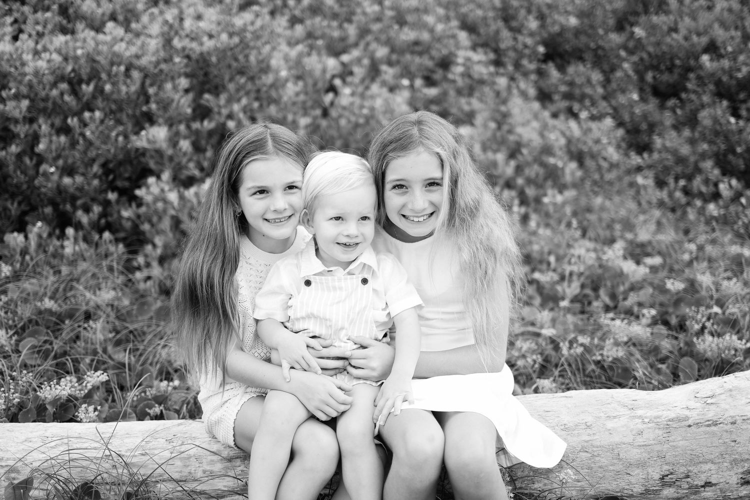 Black and white photo of three young children sitting on a log outdoors, two girls flanking a toddler boy.