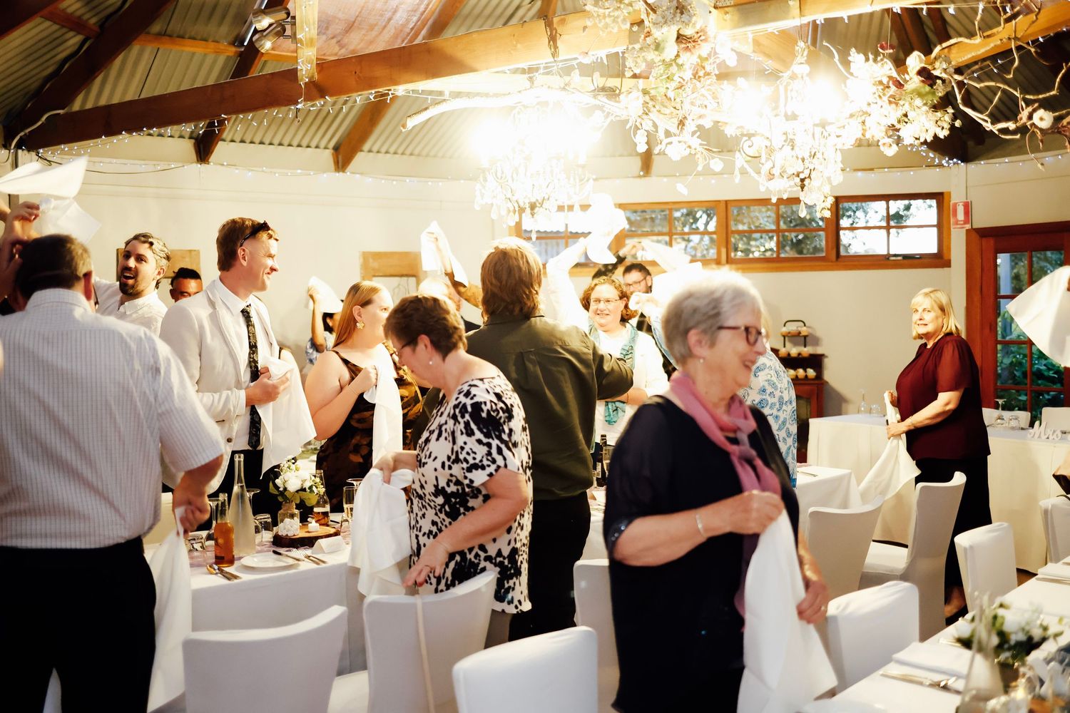 Wedding reception guests mingling in elegant venue with white chairs, chandeliers, and festive table settings.