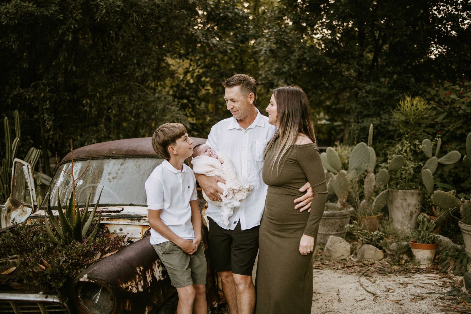 A family admires their newborn baby beside a vintage truck with cactus and lush greenery backdrop.