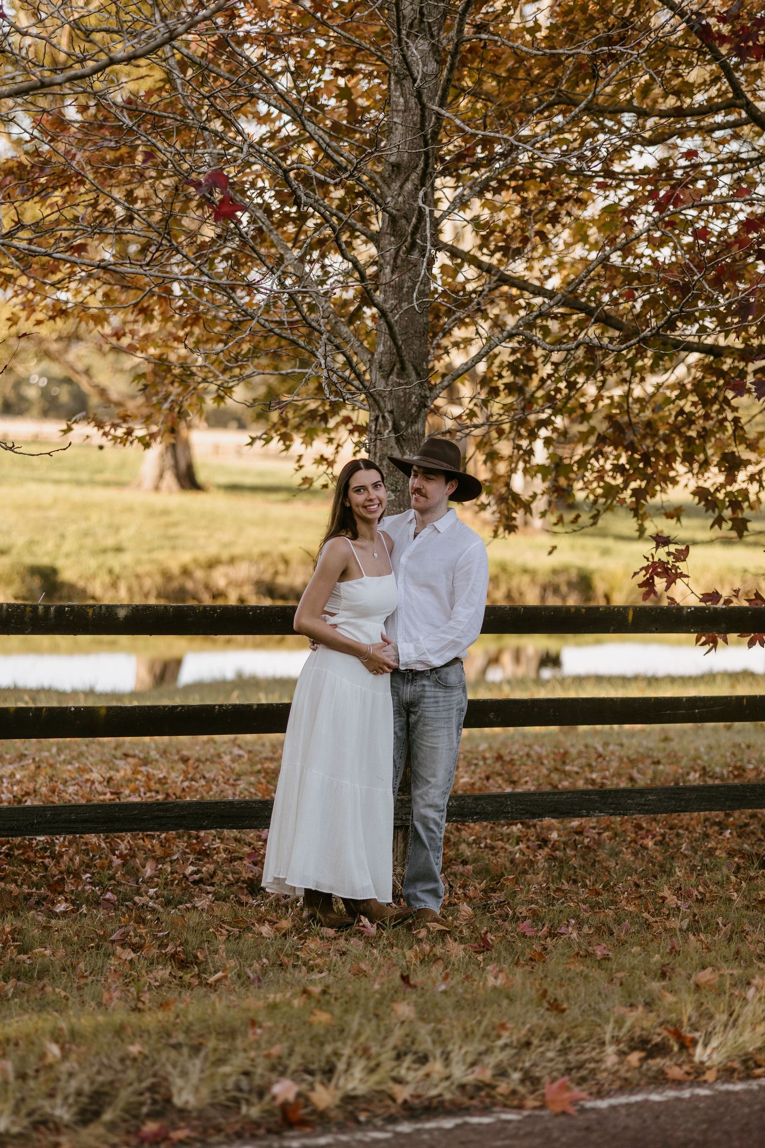 Couple embracing under autumn maple tree beside wooden fence in fall foliage setting.