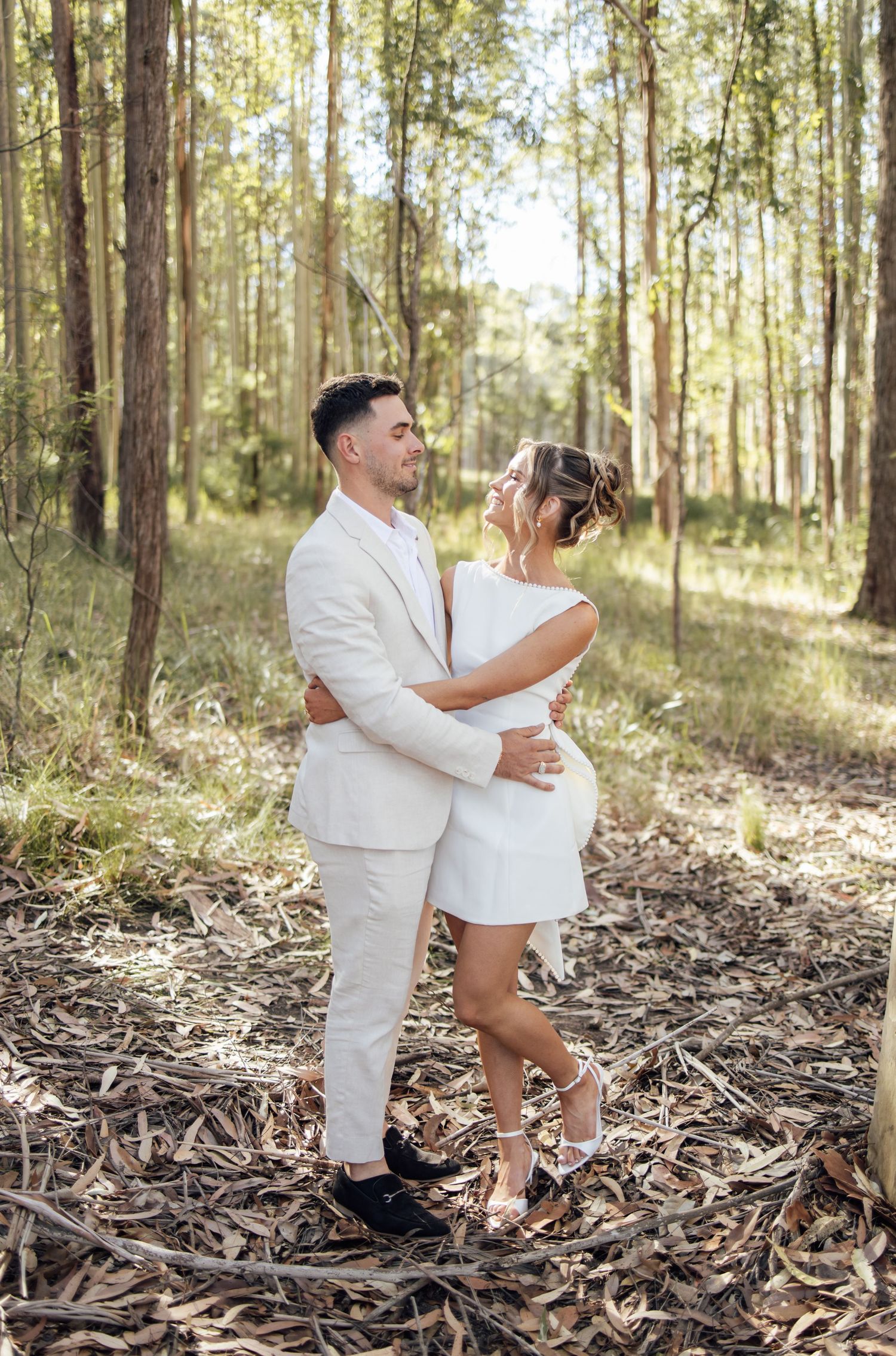 Couple in white outfits embracing in a sunlit forest during an engagement photo session.