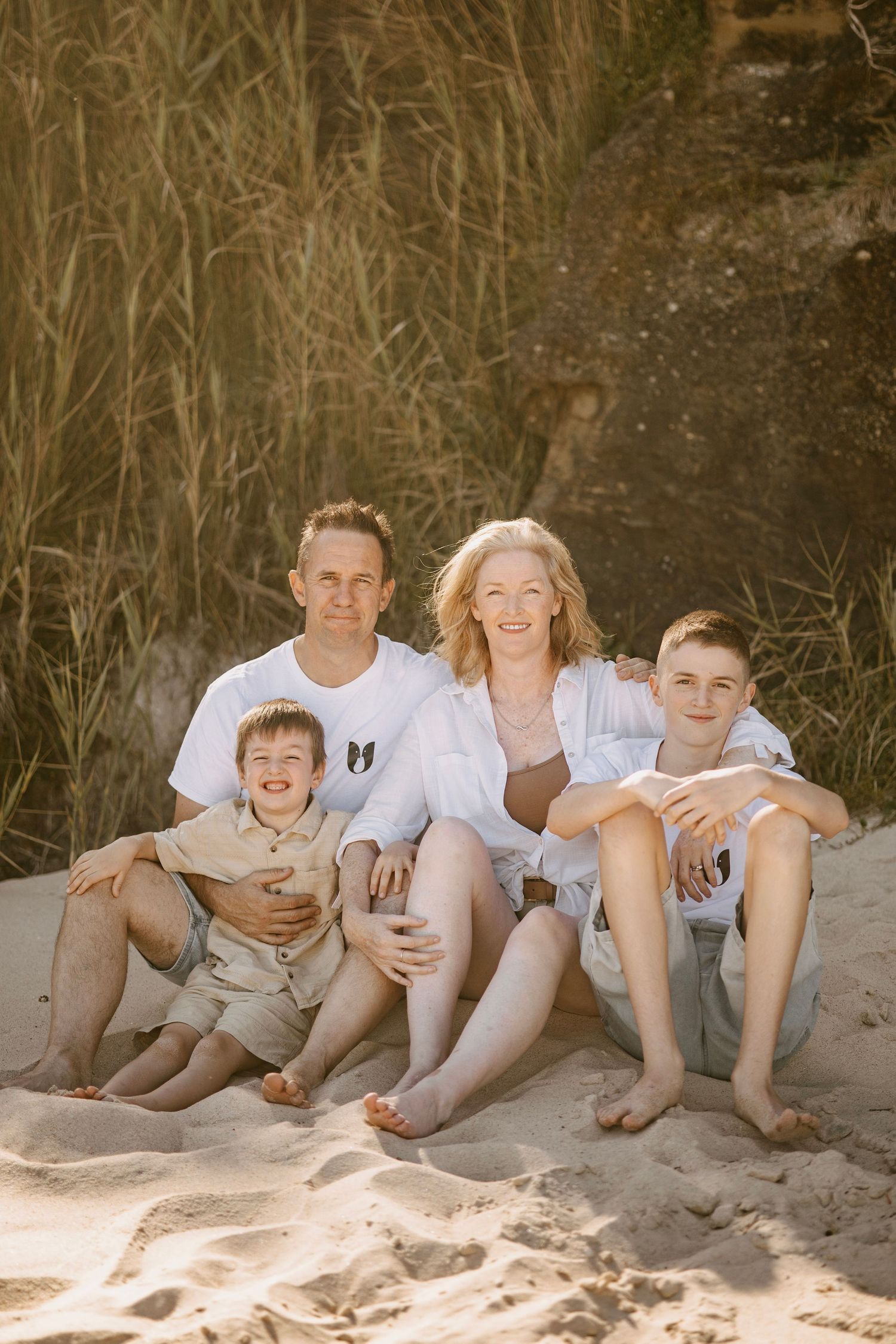 Family of four smiling together on sandy beach surrounded by tall golden grass.