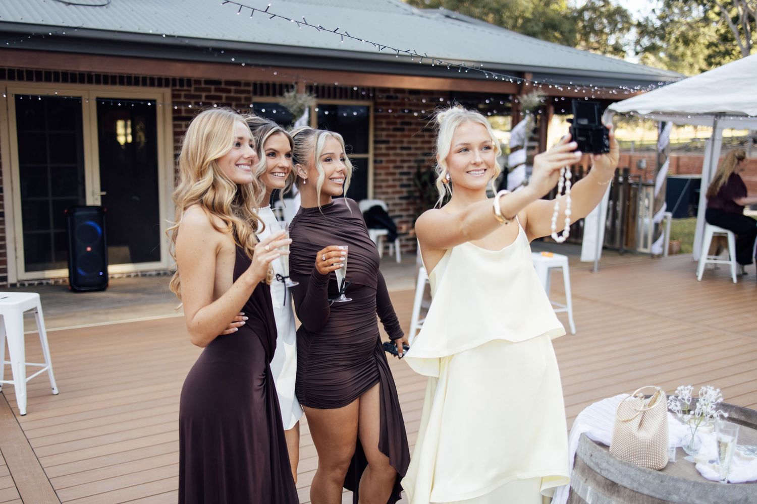 Four women in elegant dresses taking a selfie at an outdoor engagement party celebration.