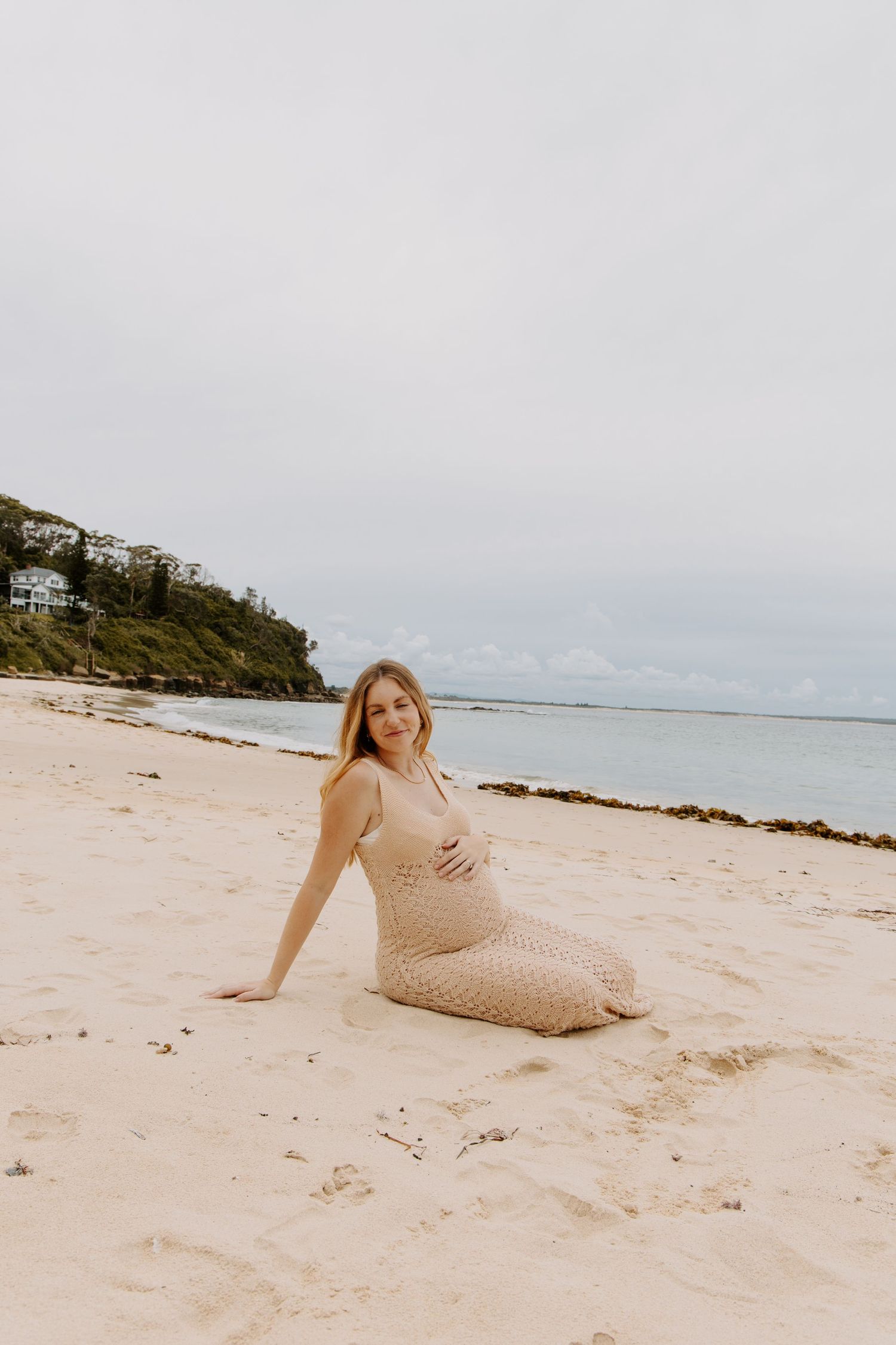 Pregnant woman in beige crochet dress sitting on white sand beach with calm ocean in background.