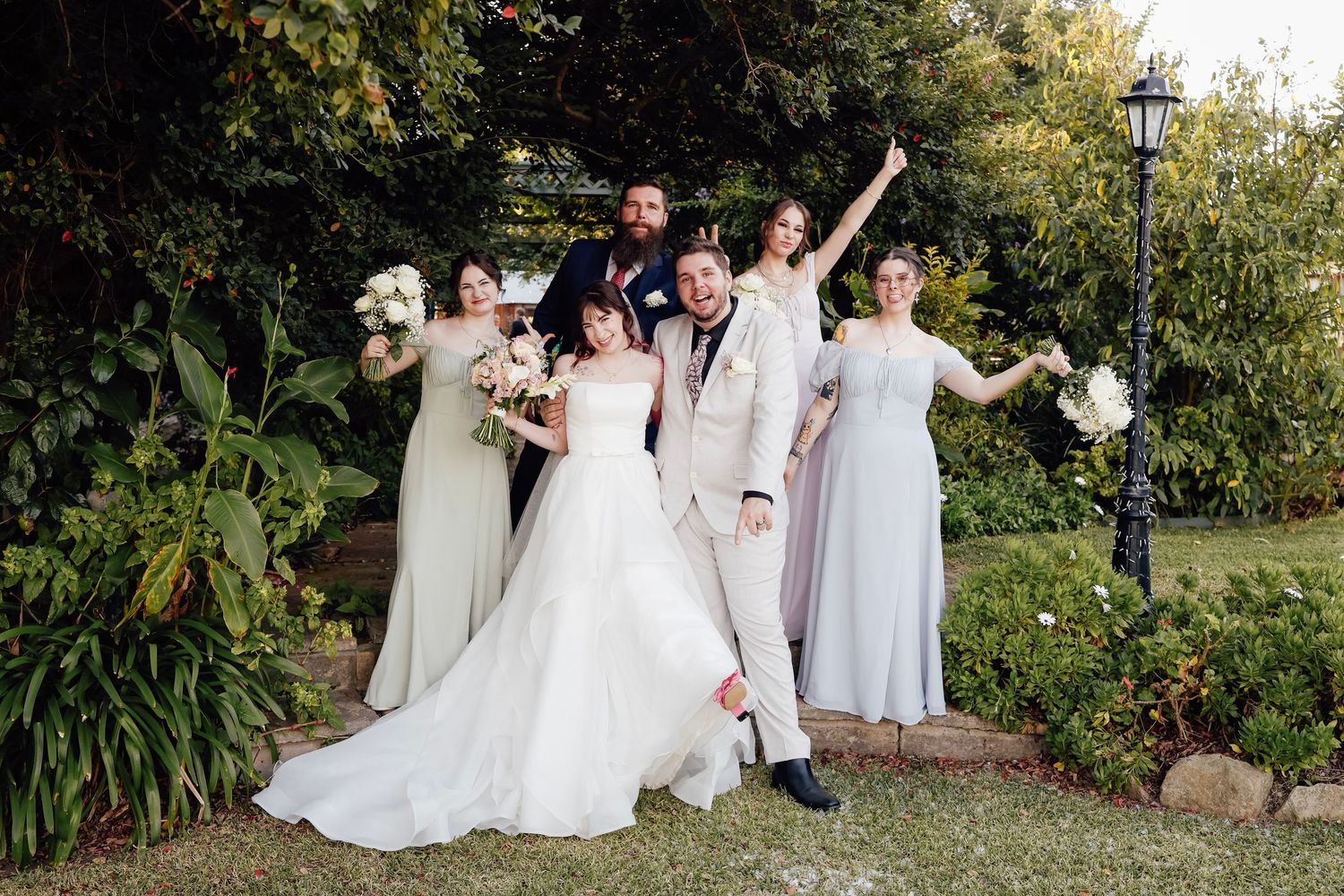 Wedding party poses joyfully in lush garden, bride in white gown, groom in tan suit, bridesmaids in sage dresses.