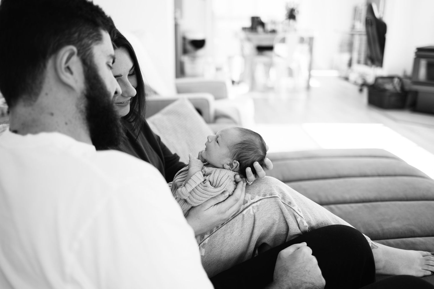 Black and white photo of parents lovingly gazing at their newborn baby on a couch at home.