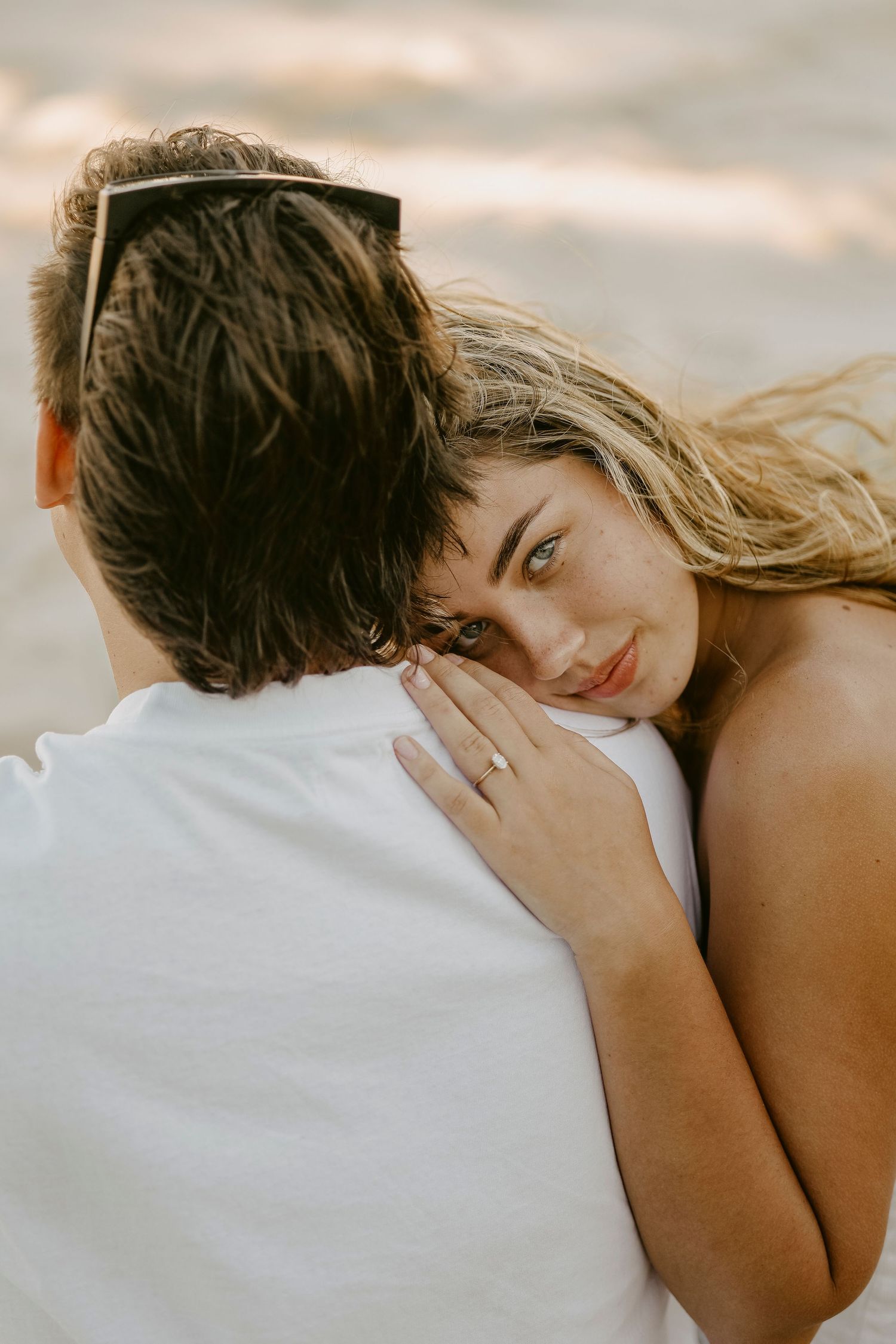 Blonde woman resting head on mans shoulder at beach, showing engagement ring while embracing closely.