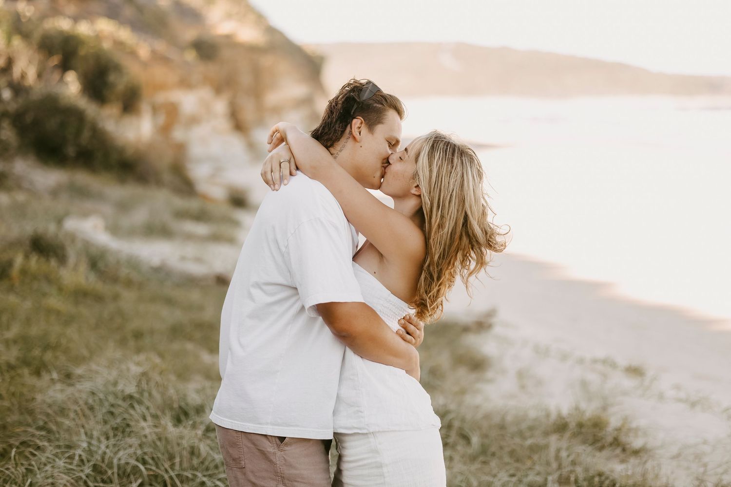 Couple sharing a romantic kiss on a scenic beach at golden hour, dressed in white outfits.